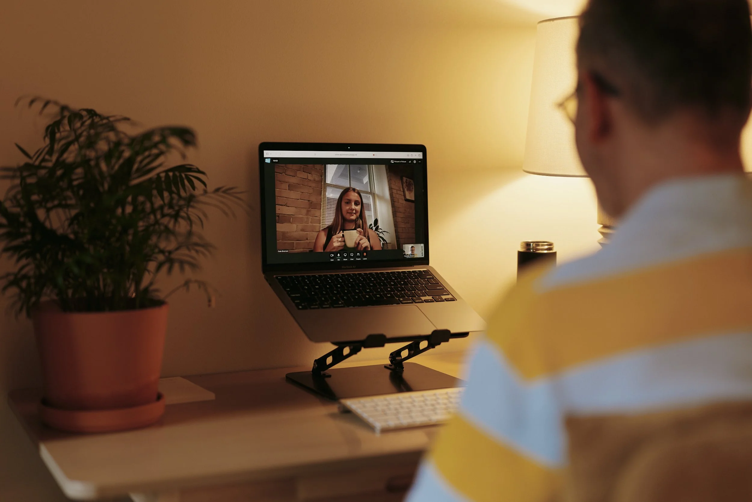 A man in a yellow striped shirt looks a computer screen where is a woman is smiling while holding a ceramic mug.