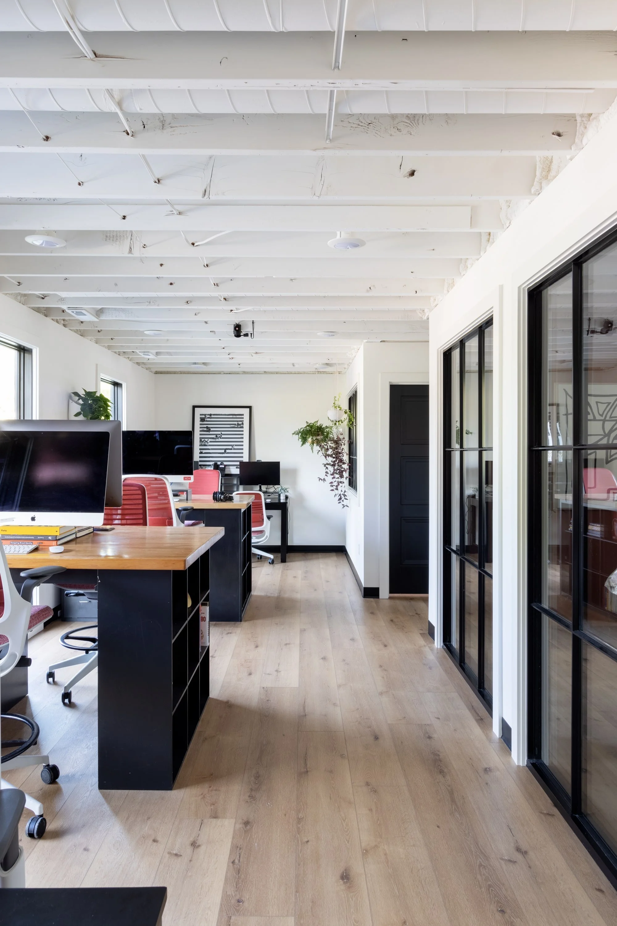 Modern open office space with wooden flooring, white ceiling with exposed beams, black-framed glass sliding doors on the right, and workstations with computers and red chairs. Potted plants are near the back wall.