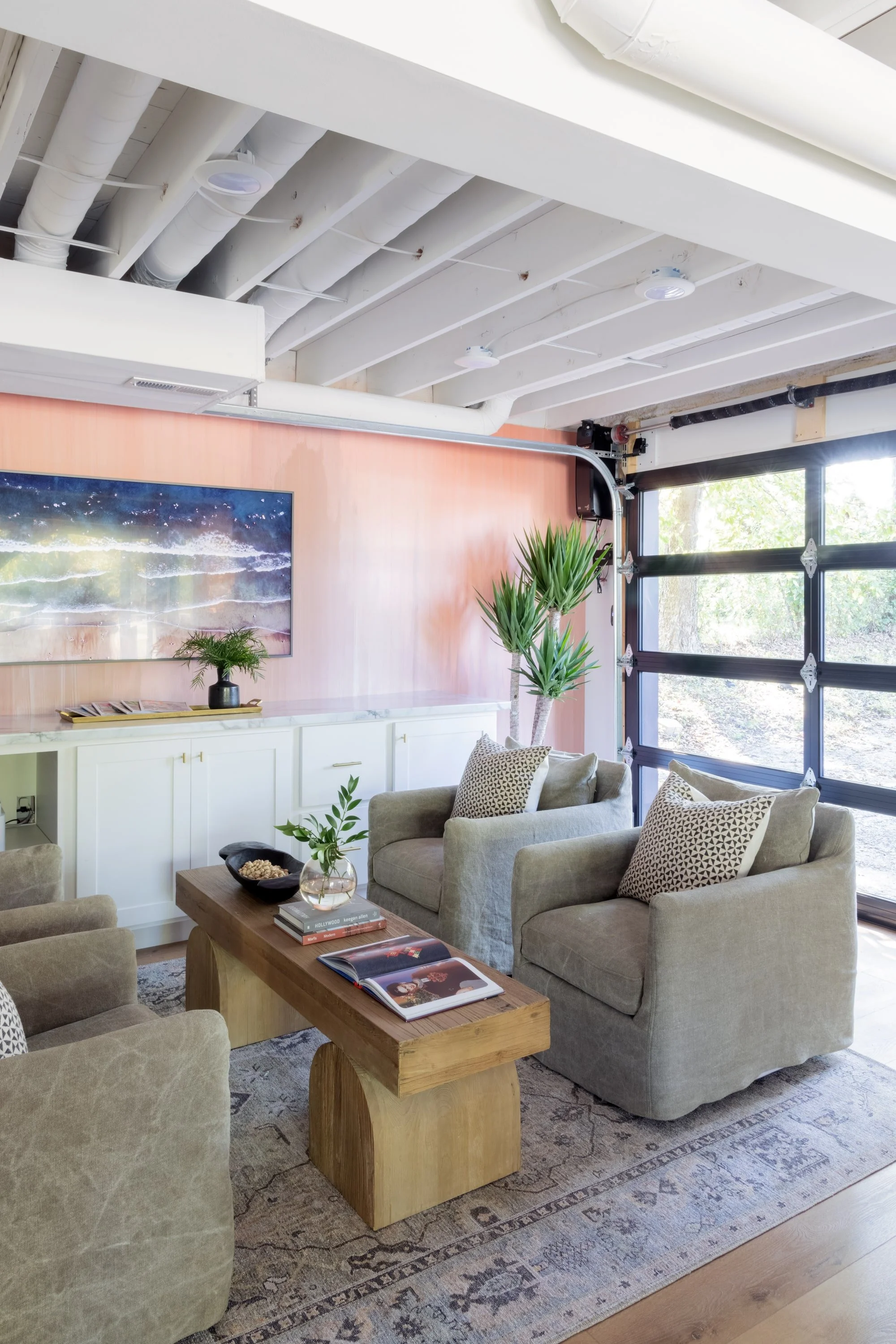 Modern living room with beige armchairs, a wooden coffee table with books, a patterned rug, a large flat-screen TV on a pink wall, plants, and a large garage door with windows, exposed ceiling pipes.