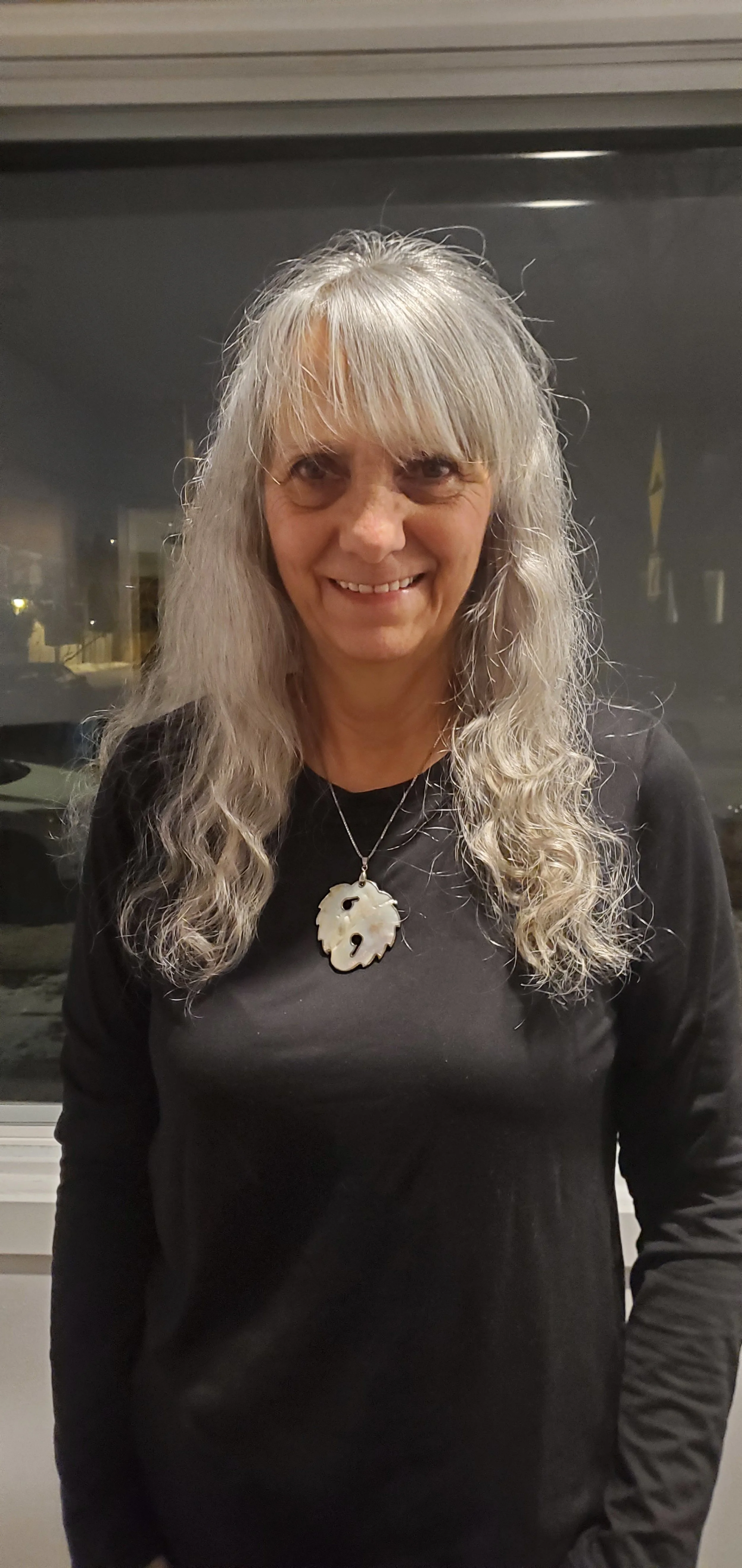 Une femme aux cheveux gris longs, portant un collier avec un pendentif en forme de visage, souriant à la caméra, vêtue d'un haut noir, devant une fenêtre la nuit.