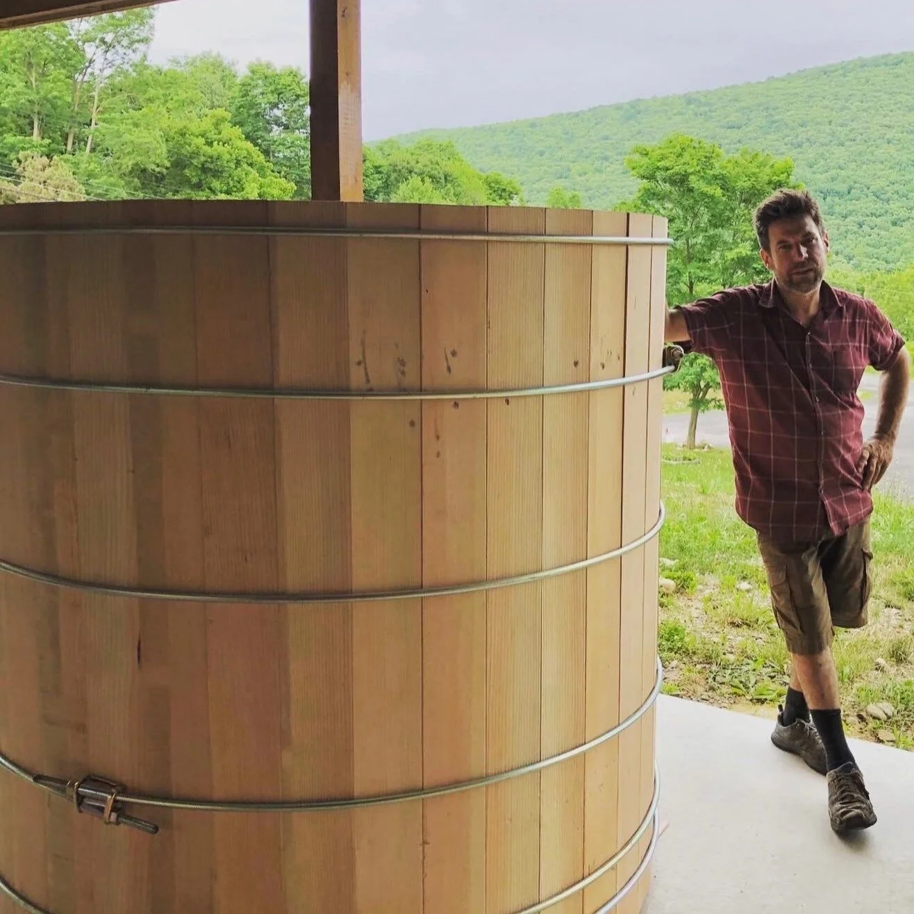 John Cox, founder of Hudson Hot Tubs, standing next to a large cedar hot tub on a porch, with a green mountain landscape and cloudy sky in the background.