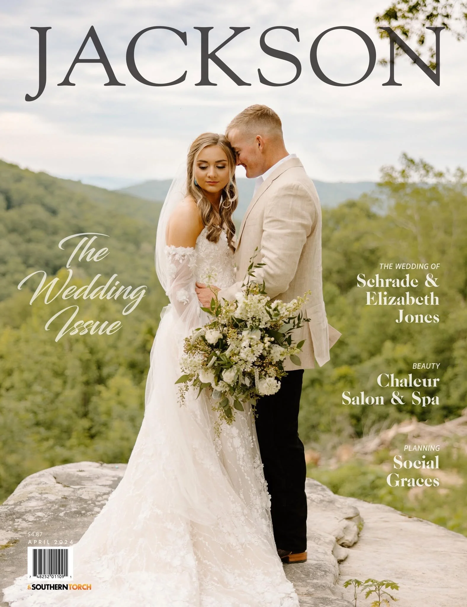 A bride and groom standing outdoors on a rocky surface, with the bride in a white lace wedding gown holding a bouquet, and the groom in a beige suit leaning their foreheads together, surrounded by greenery and a cloudy sky.
