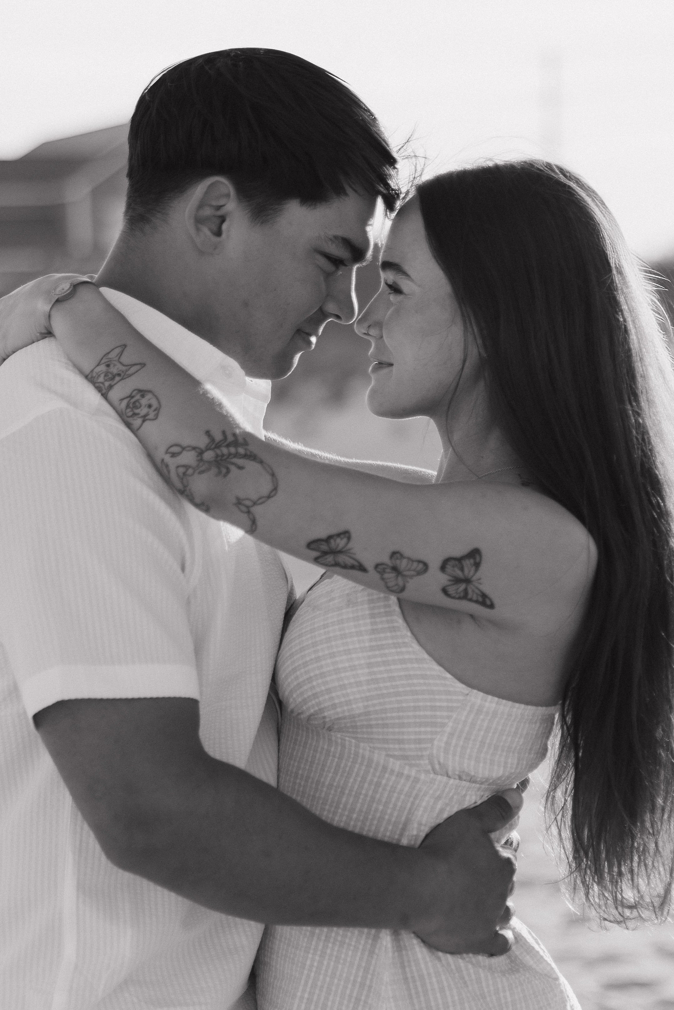 A black and white photo of a couple embracing, with their foreheads touching and eyes closed, smiling gently, outdoors.