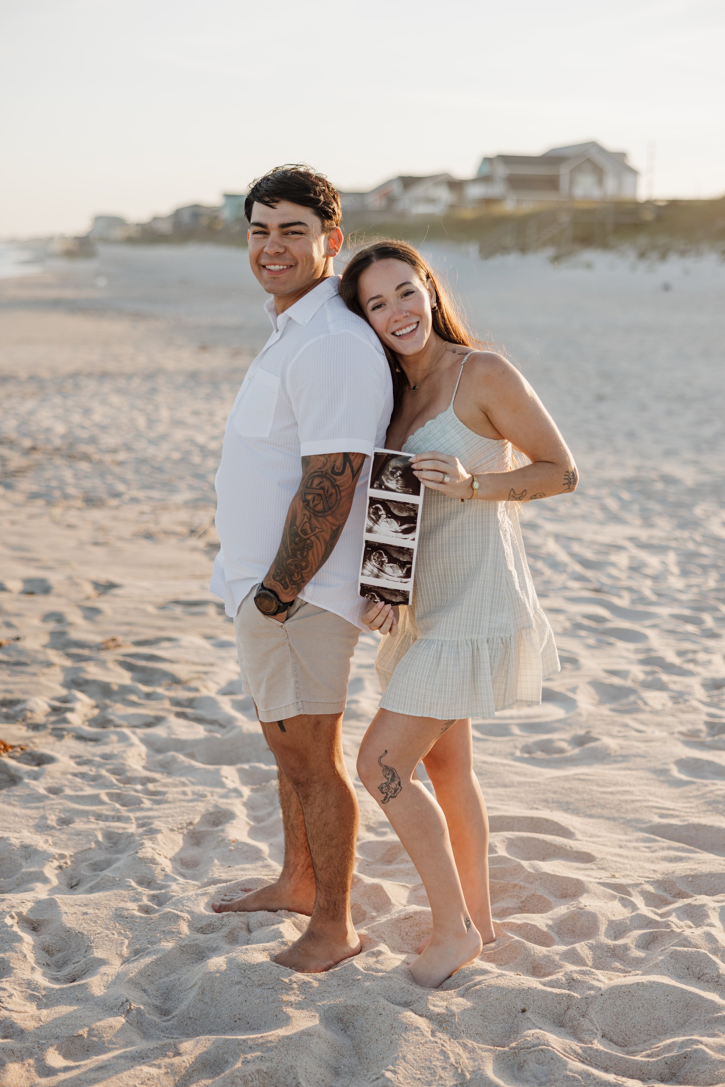 Pregnancy announcement photos on North Topsail Beach, NC couple standing on the sand with beach houses in the background