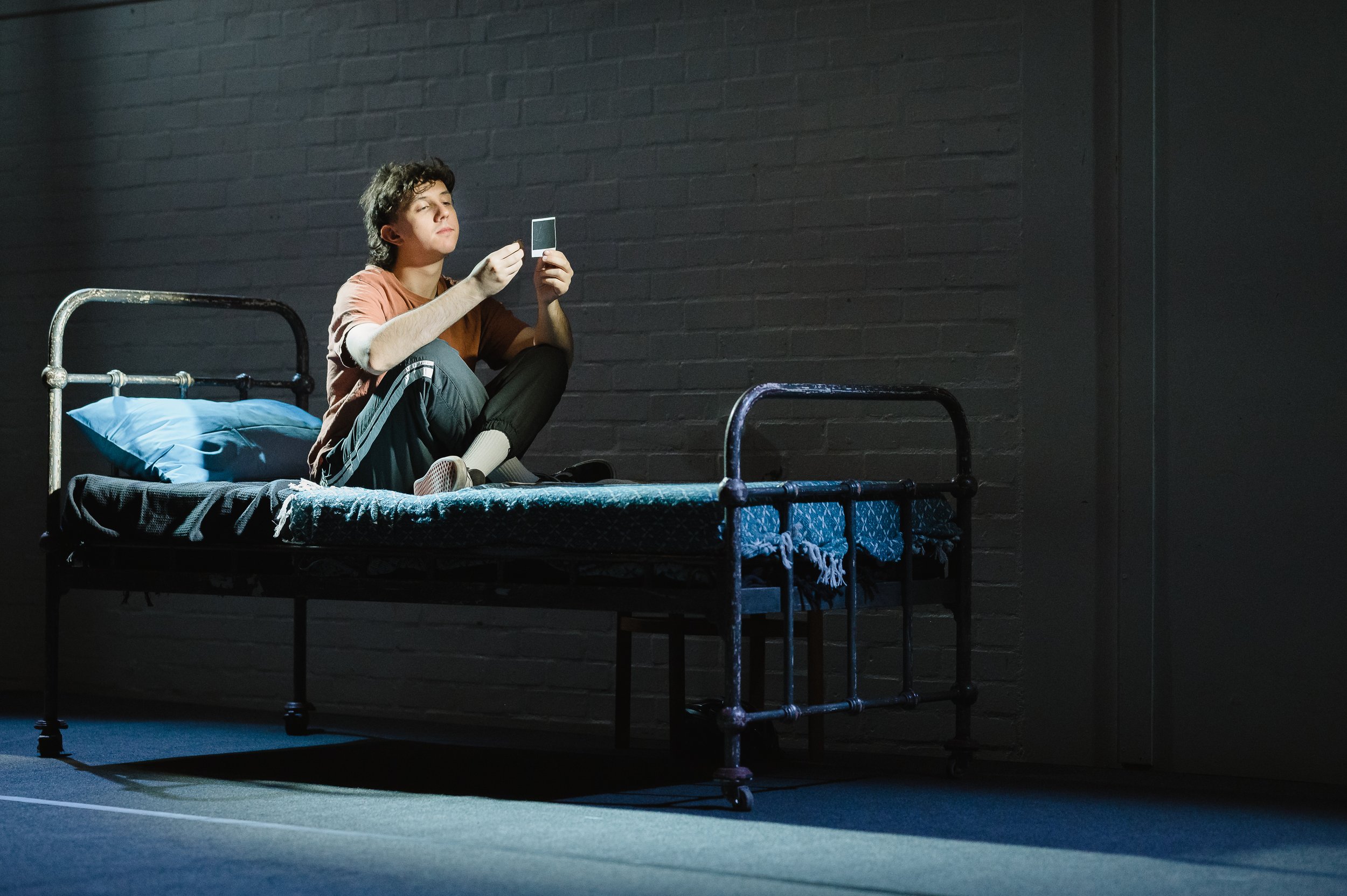 Image © Emma Hare. An actor sits on a bare bed on a stage in a pool of light. Image from Teulu