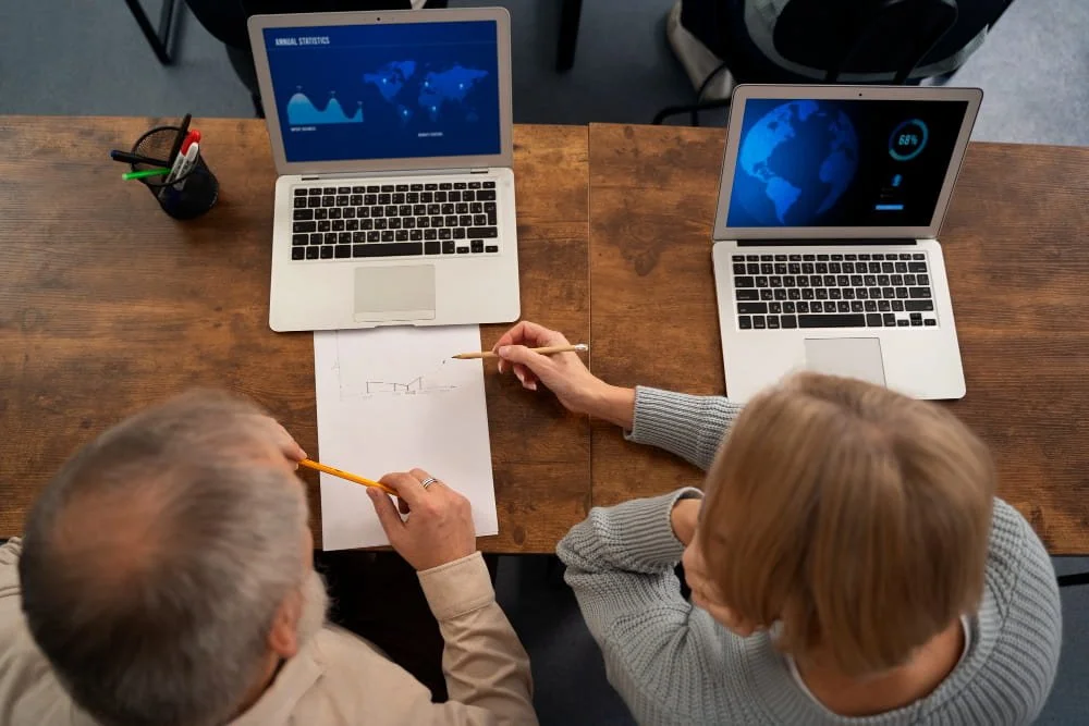 Two people discussing at a wooden table with laptops displaying statistics.