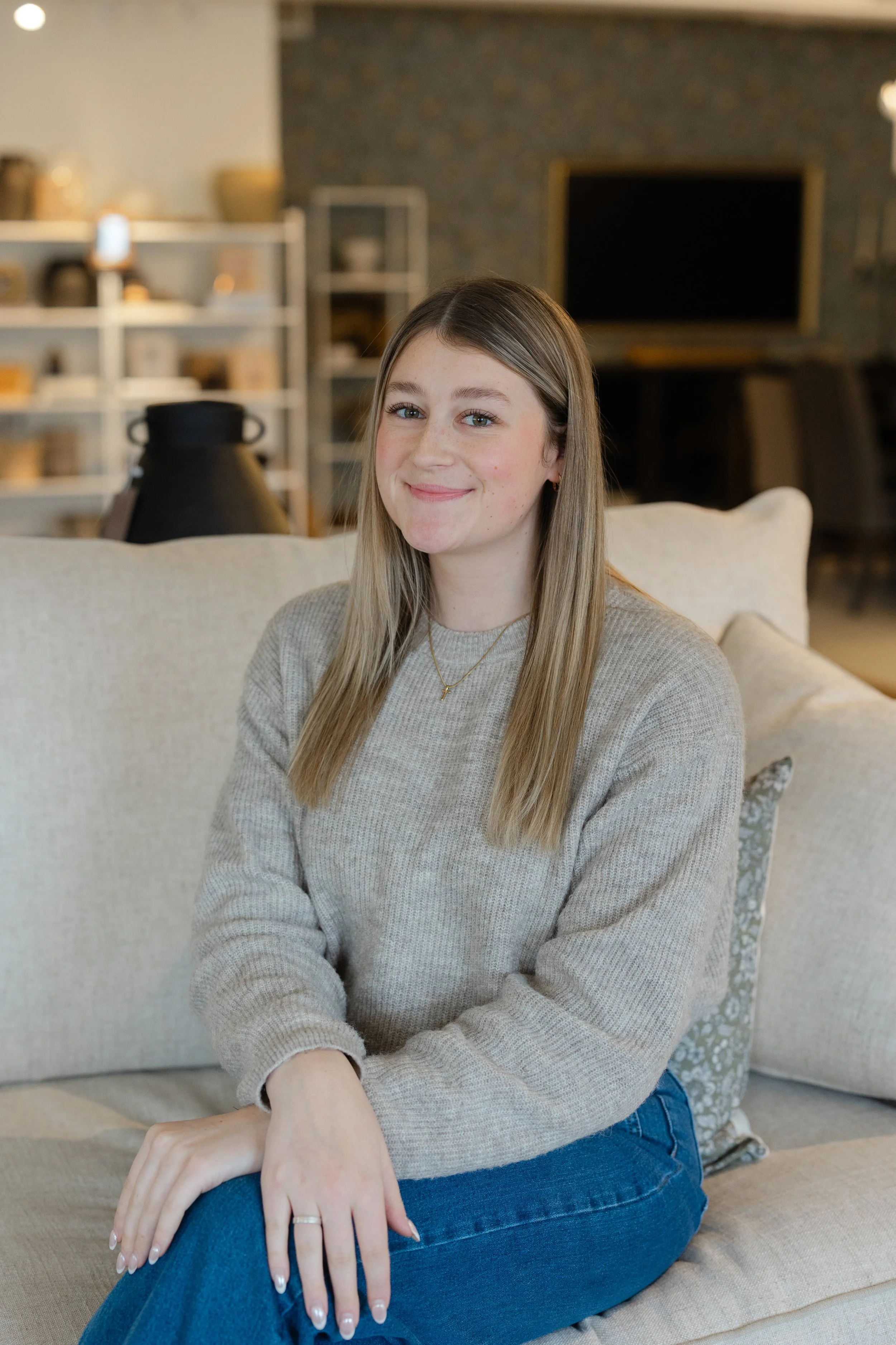 A young woman with long blonde hair sitting on a beige sofa in a cozy living room, wearing a beige sweater and blue jeans, smiling softly at the camera.