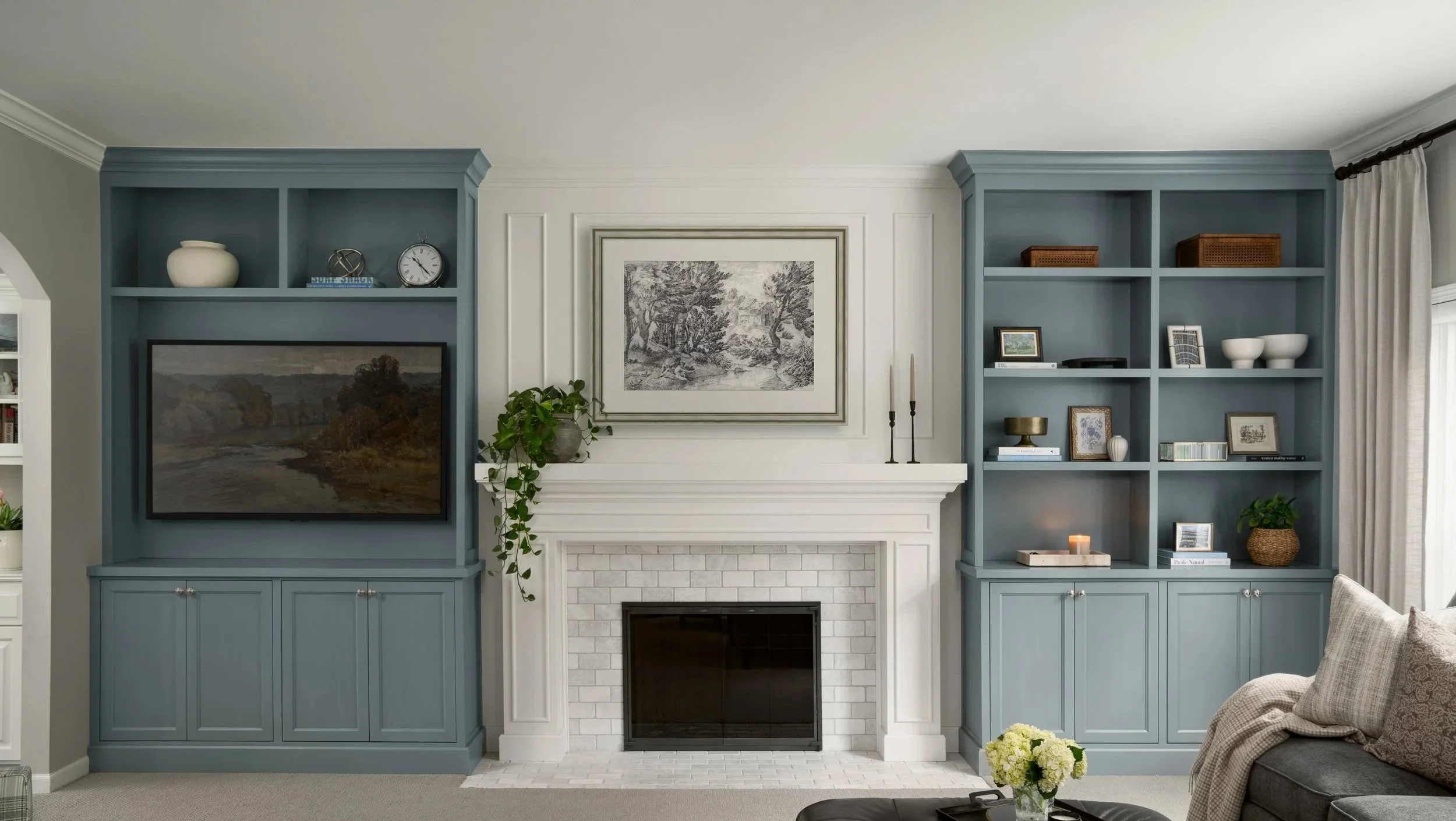 Living room with a white brick fireplace, blue built-in shelves on both sides, and a framed black-and-white landscape drawing above the mantel. Decor includes a potted plant, candles, framed photos, vases, and baskets, with curtains partially visible on the right.