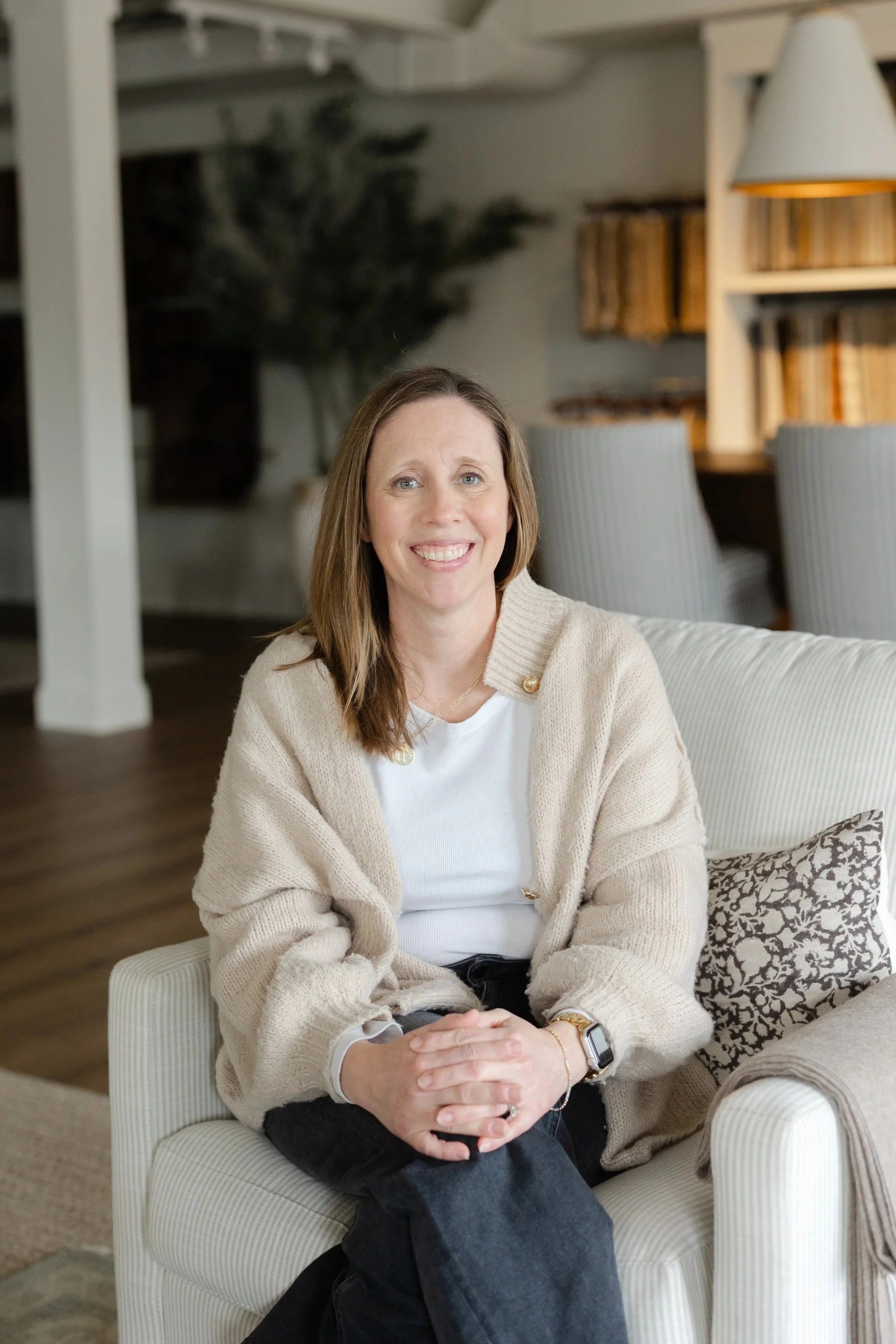 A woman with shoulder-length brown hair smiling, seated on a light-colored striped armchair in a cozy living room with a bookshelf and a potted plant in the background.