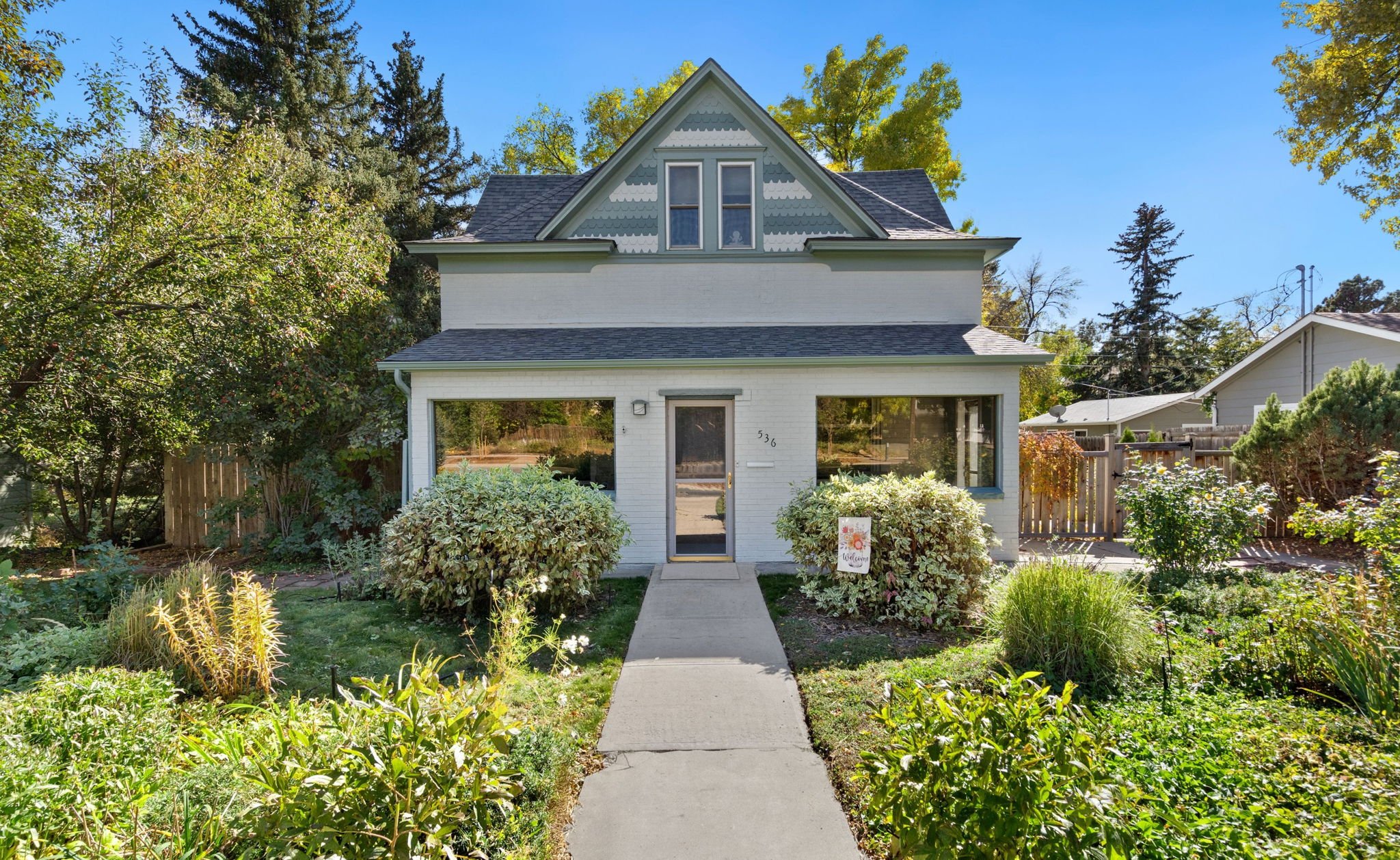 Front view of a two-story house with a steep gabled roof, large windows, and a welcoming front yard garden with various shrubs and plants. The house has a light-colored exterior and is surrounded by mature trees under a clear blue sky.