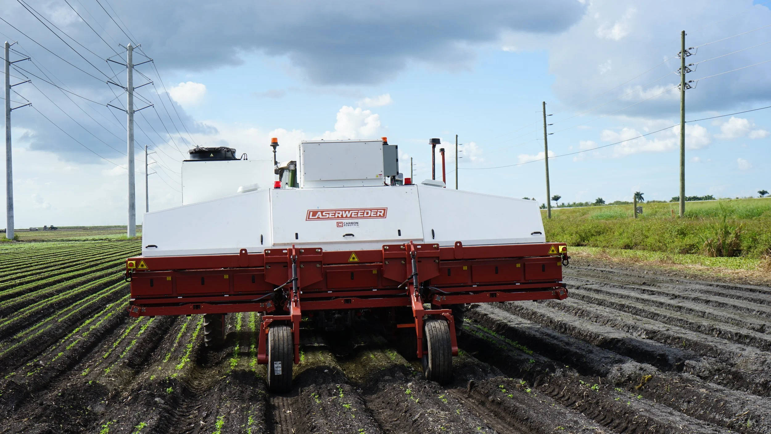 A Laserweeder machine operating in a farm field with rows of crops and power lines in the background.