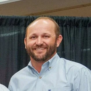 Smiling man in a blue shirt standing in front of a black curtain.