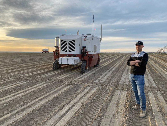 Hombre de pie en un campo con un robot agrícola en una granja al atardecer.