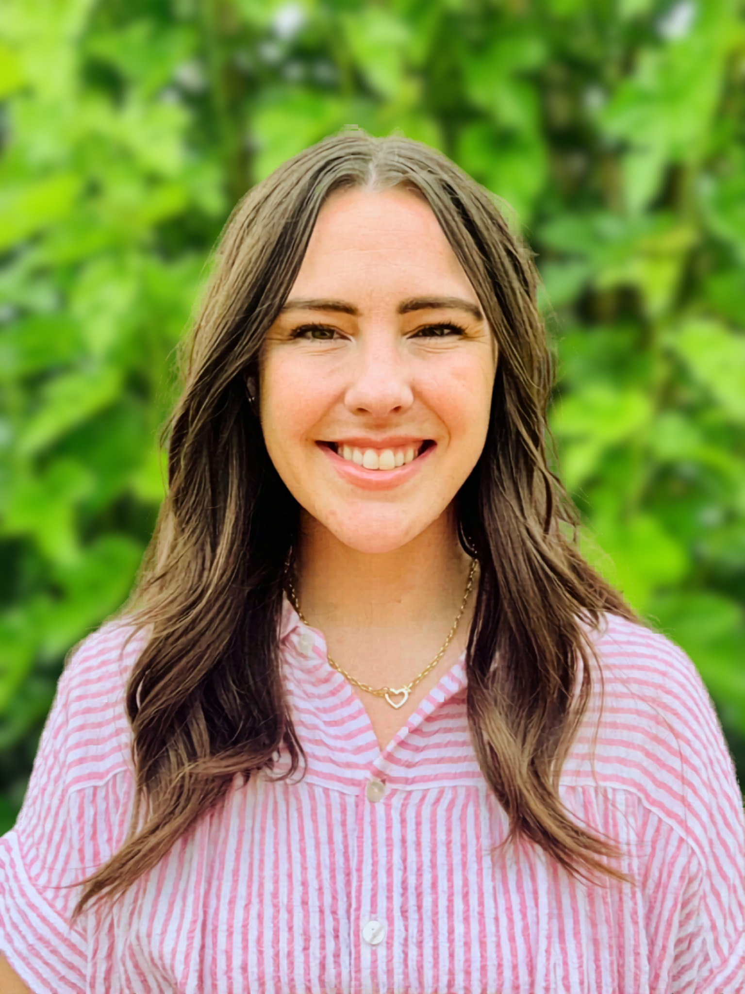A woman with long brown hair wearing a pink and white striped shirt, smiling, with green leafy trees in the background.