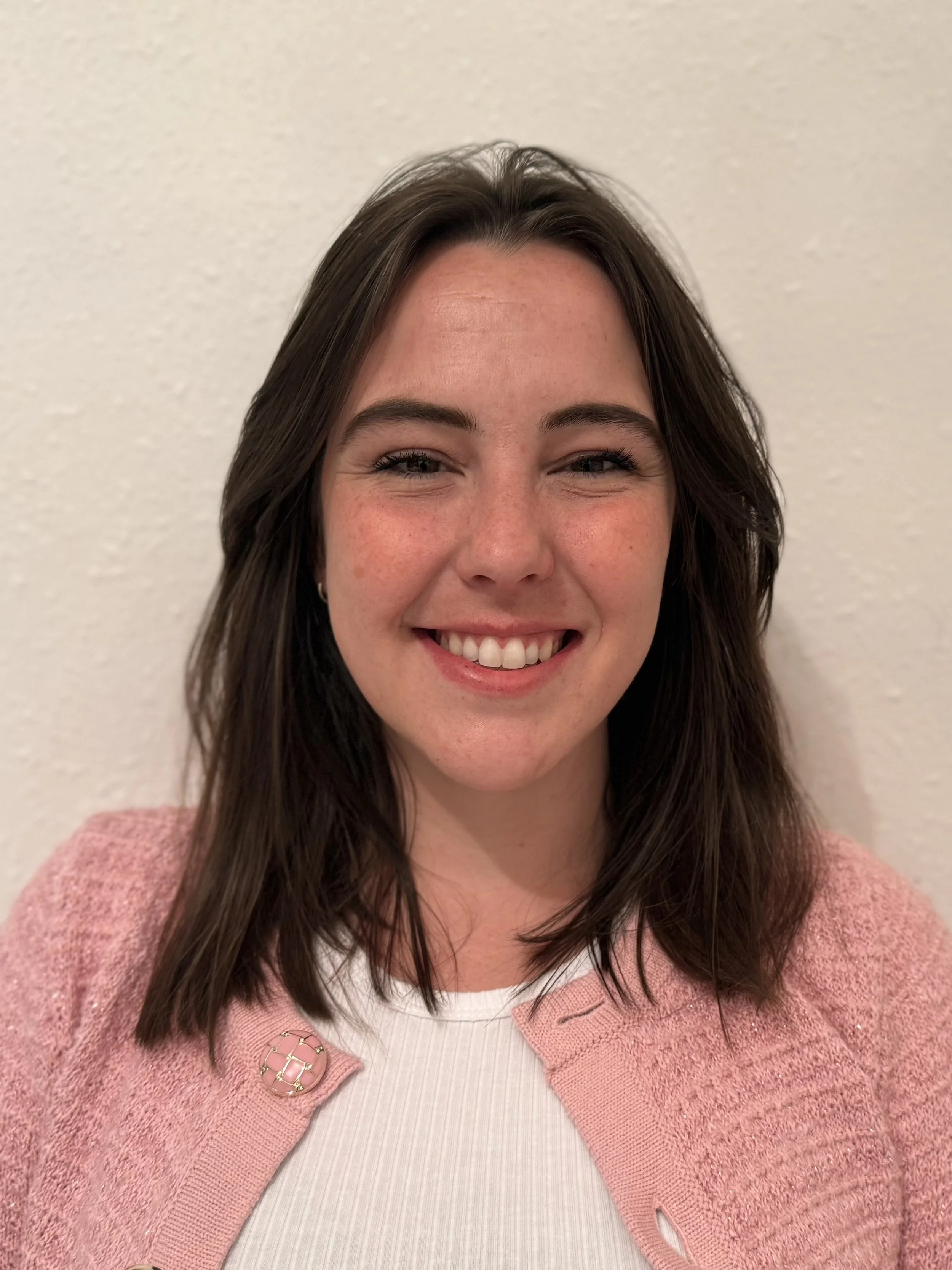Portrait of a young woman with shoulder-length brown hair, wearing a pink cardigan and a white top, smiling against a neutral background.