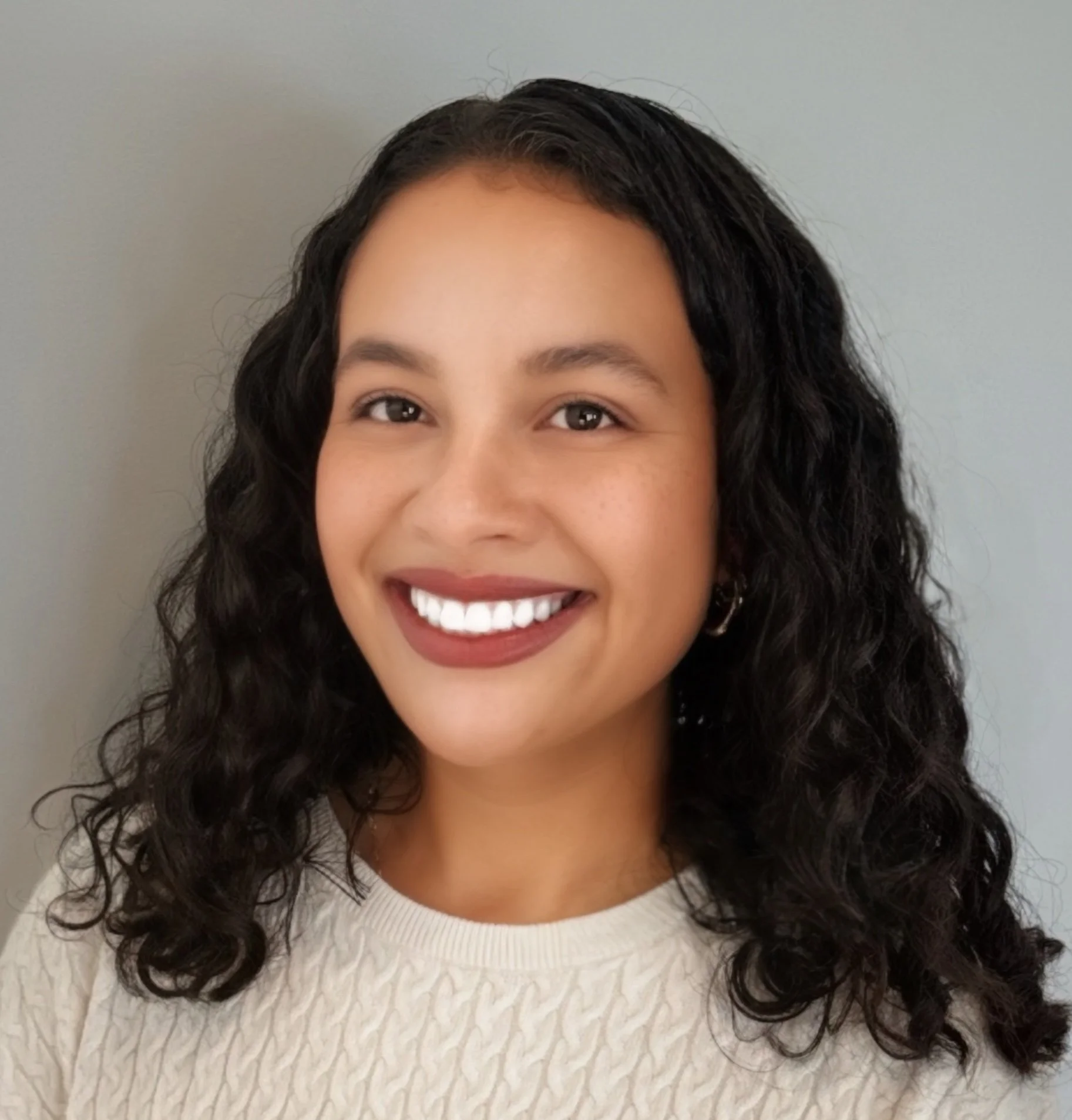 A young woman with curly dark hair, smiling, wearing a cream textured sweater, standing against a light gray background.