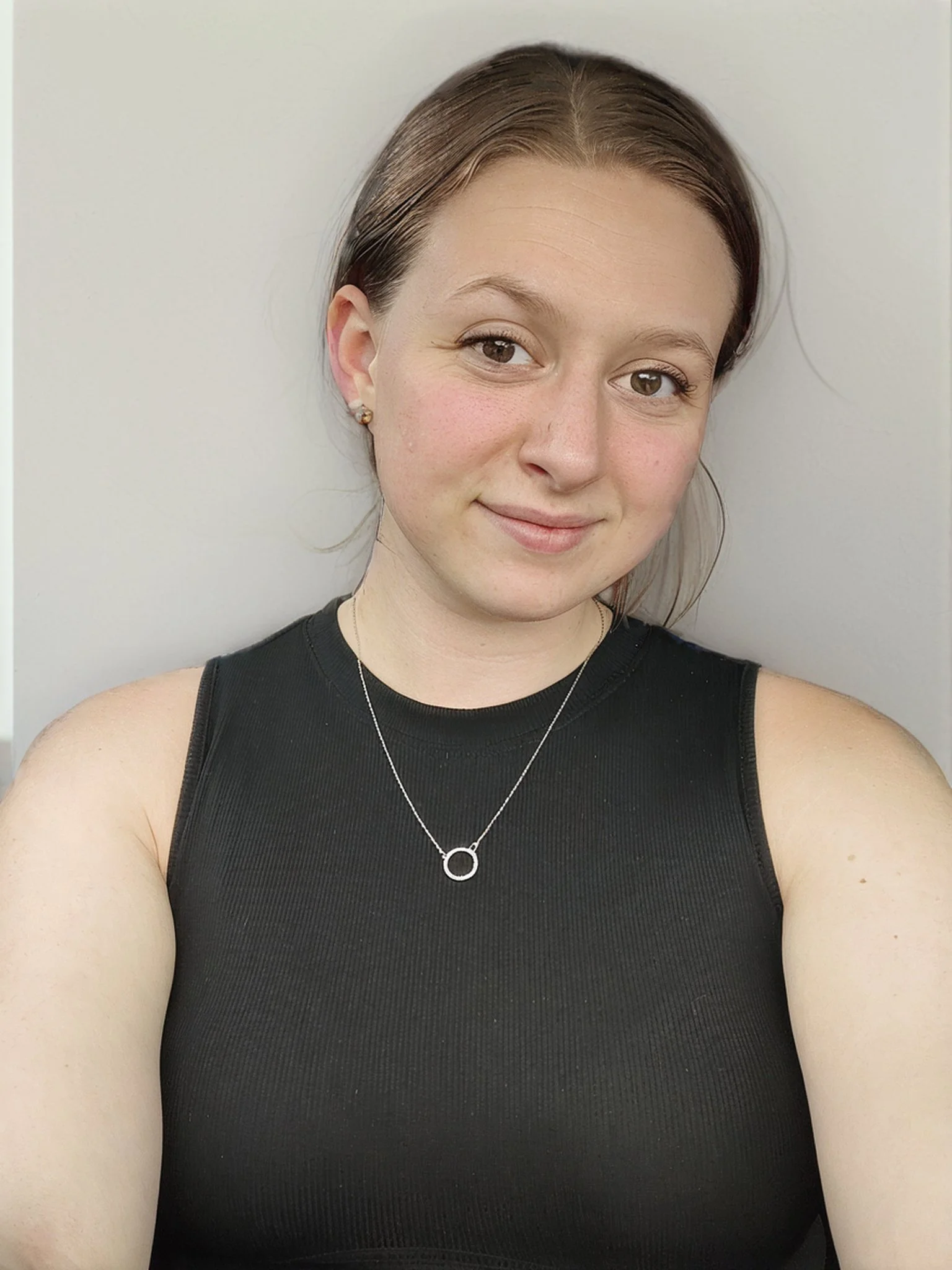 A young woman with brown hair, light skin, wearing a black sleeveless top, a silver necklace with a circular pendant, and small earrings, smiling against a neutral background.