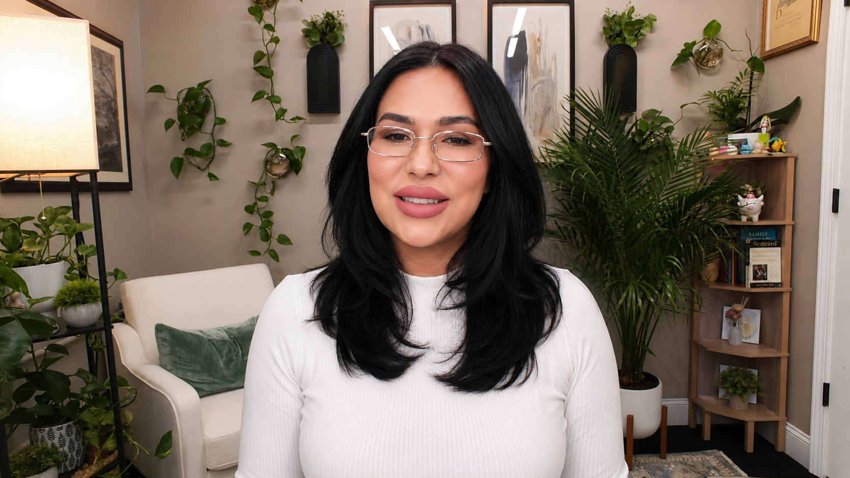 A woman with black hair, glasses, and a white top smiling in a cozy, decorated living room with plants, artwork, and books.