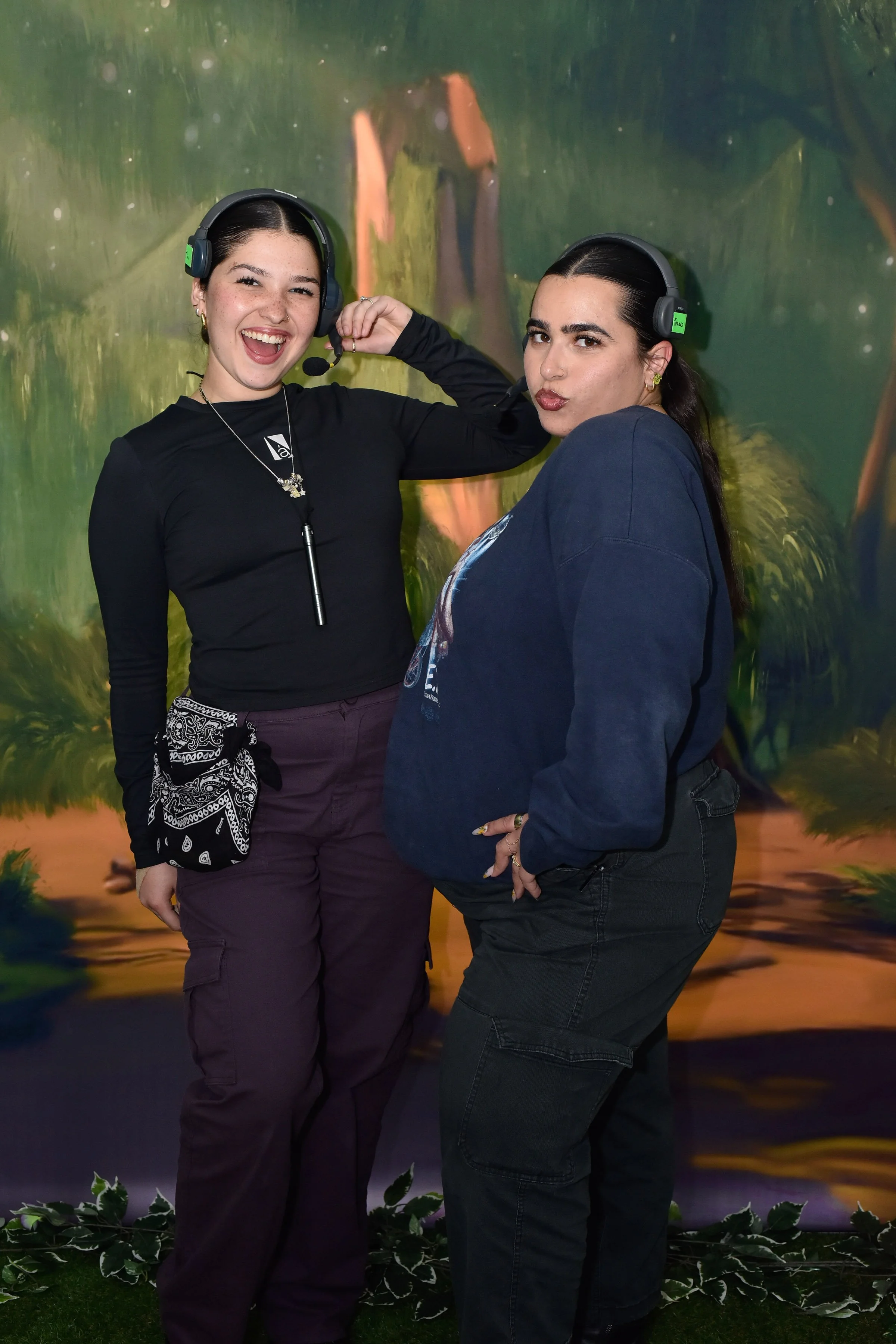Two young women wearing black headsets with microphones pose confidently in front of a colorful, nature-themed mural. One woman is smiling widely, fairy-tale style, while the other has a slight smirk, both dressed in dark clothing.