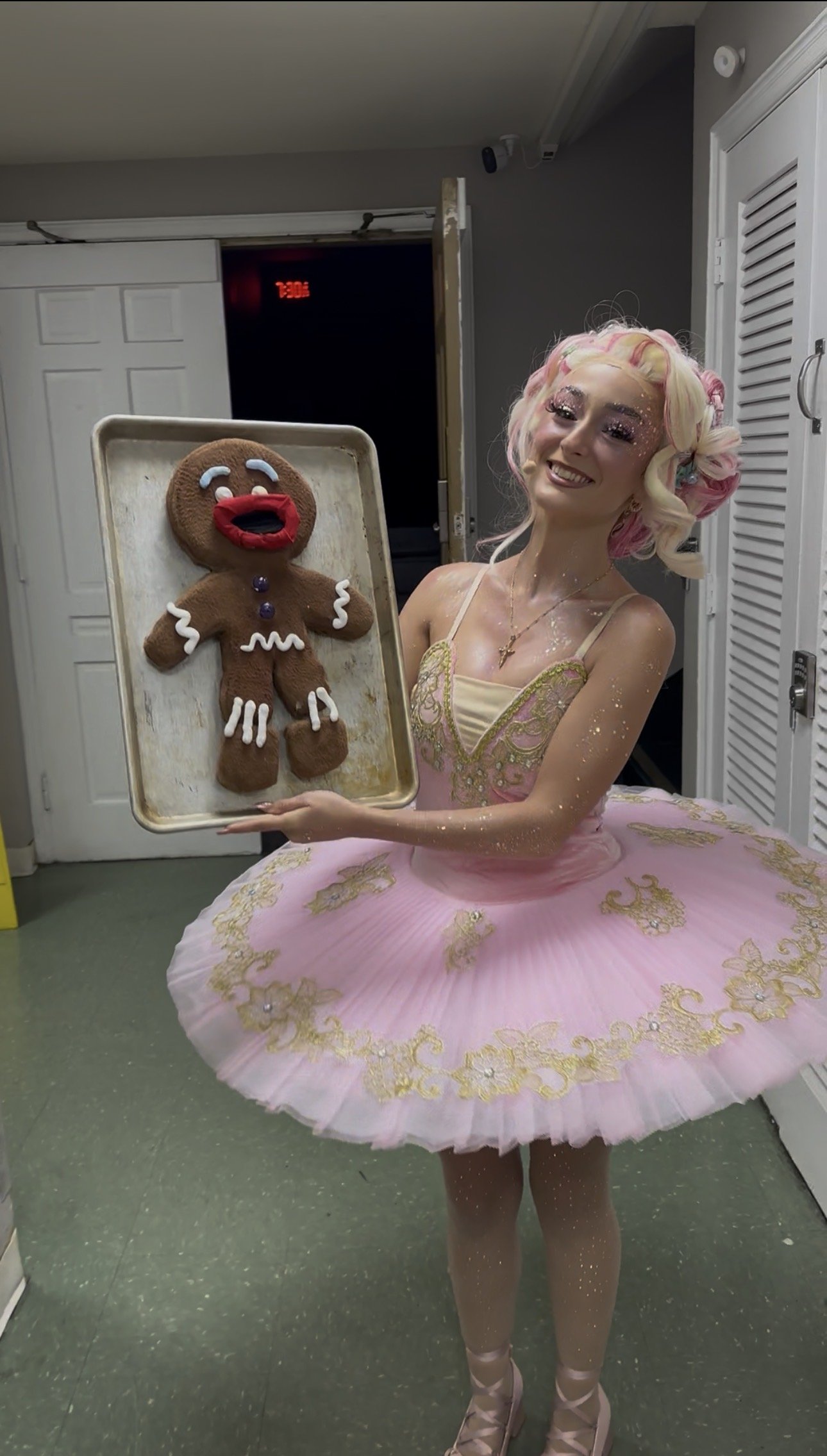 A woman dressed as a ballerina with a pink tutu and glitter on her skin, smiling and holding a tray with a gingerbread cookie shaped like a smiling face.
