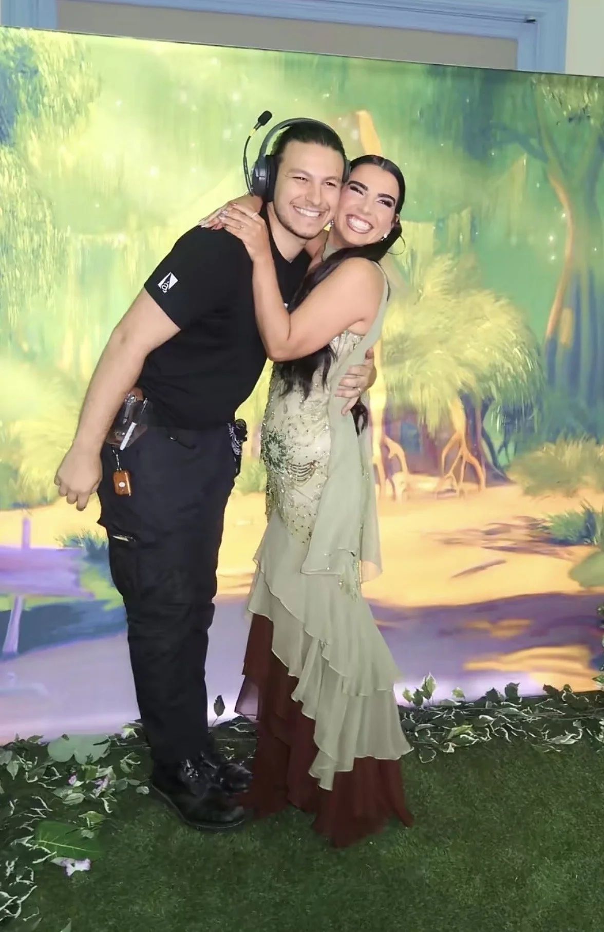 Two people, a man and a woman, are hugging and smiling in front of a colorful forest-themed backdrop. The man is wearing a black t-shirt and black pants, and the woman is wearing a beige and maroon dress with intricate embroidery. They are standing o