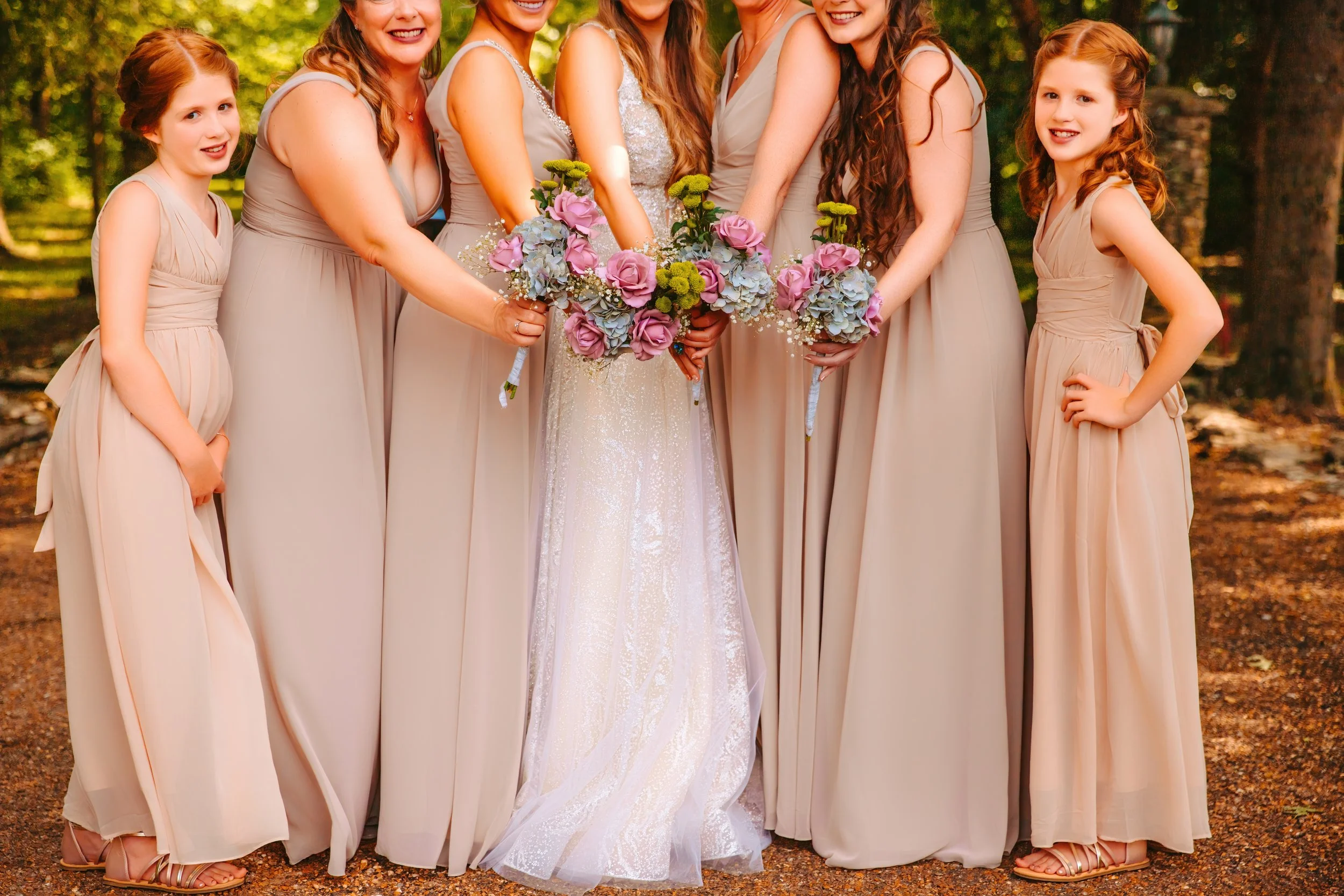 Group of seven women and two young girls in beige dresses standing outdoors, holding bouquets of pink and purple flowers, smiling with trees in the background.