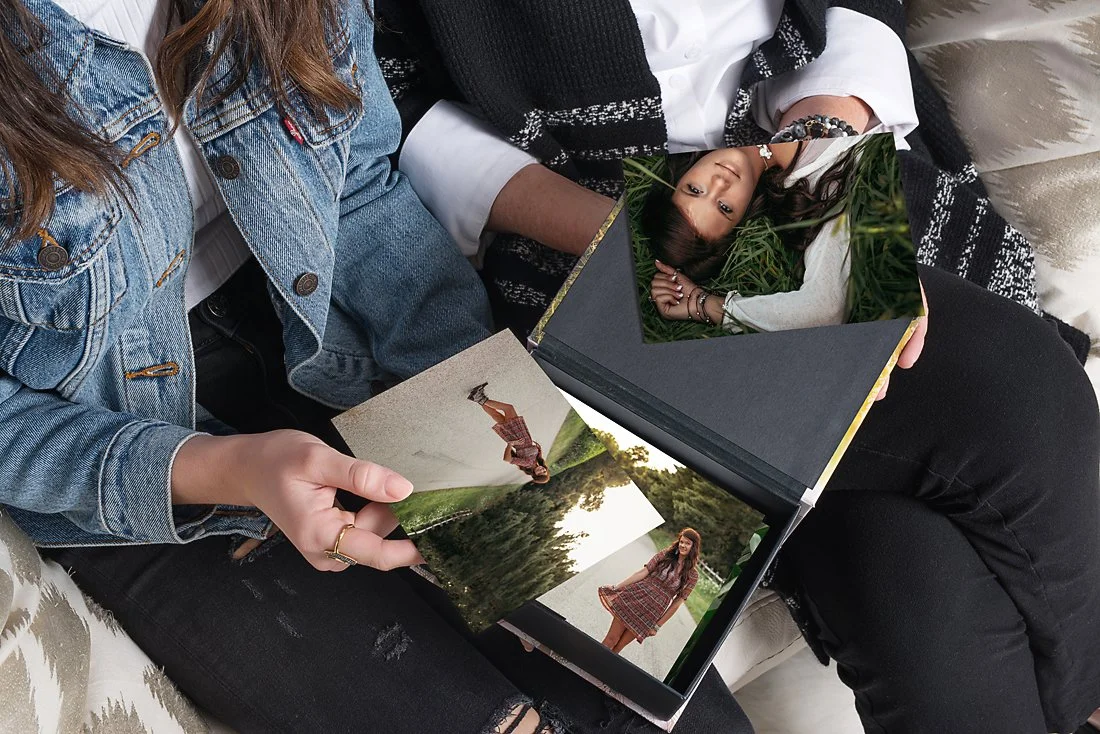 Two people sitting close together on a sofa, looking through a photo album with pictures of a woman and a child, while holding a photograph of a child doing a flip.