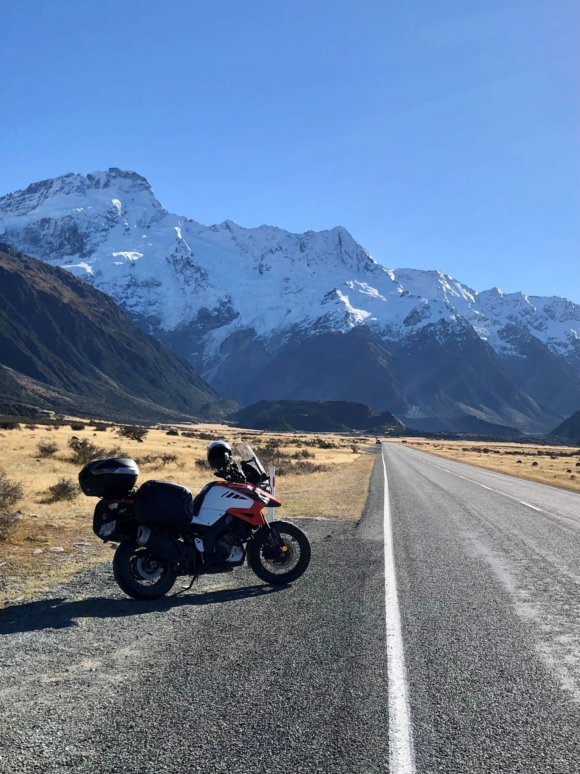 A motorcycle parked on the side of a two-lane road with mountains covered in snow in the background and a clear blue sky.