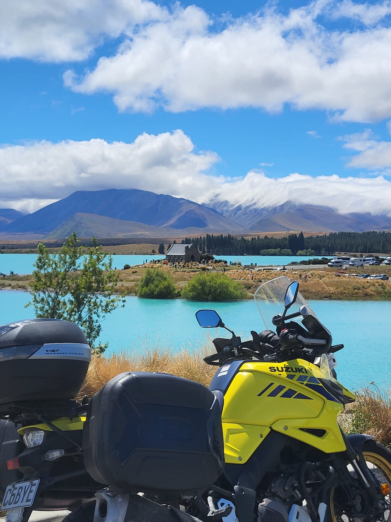Two motorcycles parked near a turquoise body of water with a mountain range and partly cloudy sky in the background.
