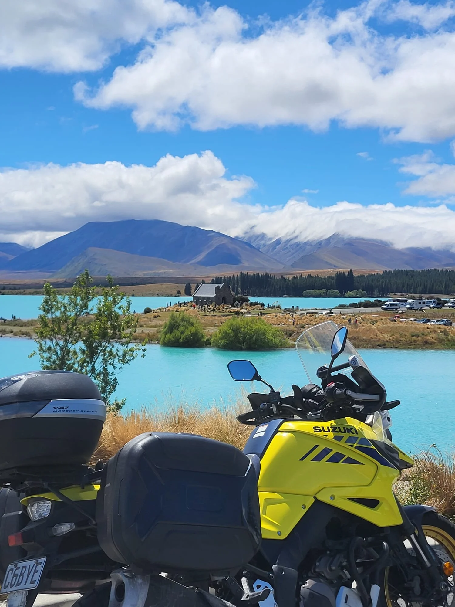 Two motorcycles parked near a turquoise body of water with a mountain range and partly cloudy sky in the background.