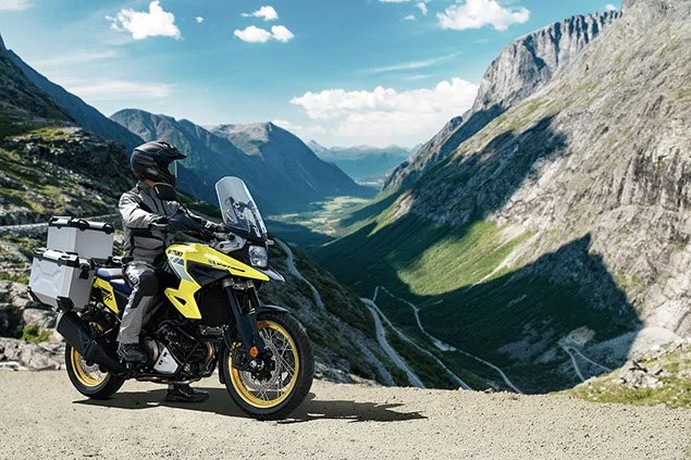 Motorcyclist riding a yellow adventure motorcycle along a mountain road with steep cliffs and a view of a deep valley with winding roads below.