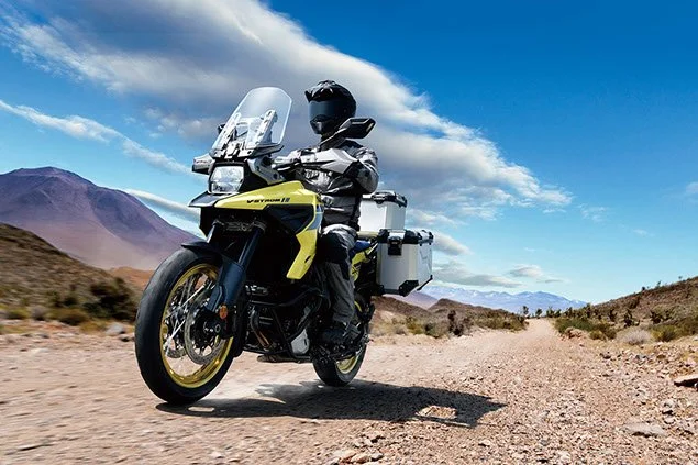 Motorcyclist riding a yellow adventure motorcycle on a dirt trail in a desert landscape with mountains and blue sky