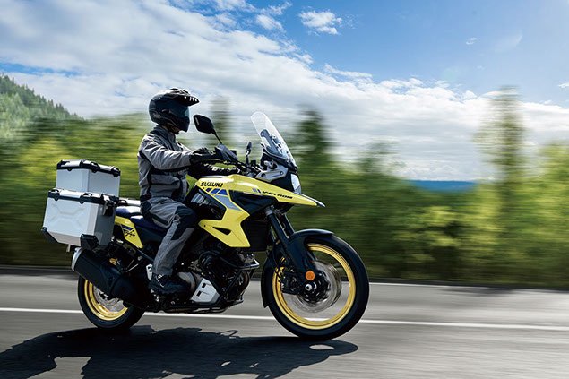 Motorcyclist riding a yellow Suzuki adventure motorcycle on a highway with green trees and a partly cloudy sky in the background.