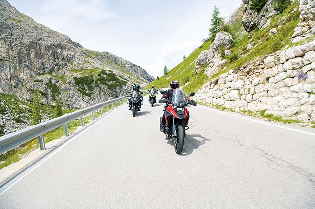 Group of motorcyclists riding on a winding mountain road surrounded by rocky cliffs and green hills.