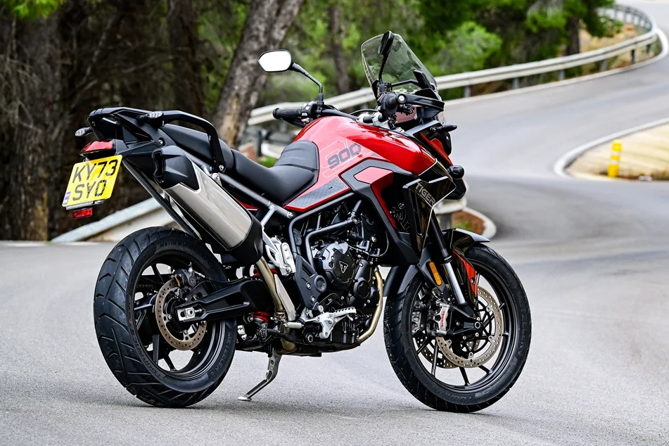 Red and black Triumph Tiger 900 motorcycle parked on a winding mountain road with trees in the background.