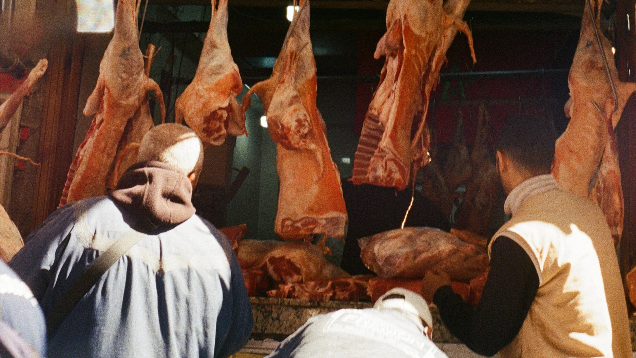 Two men with shaved heads wearing casual clothing viewing large hanging cuts of meat in a butcher shop or meat market.