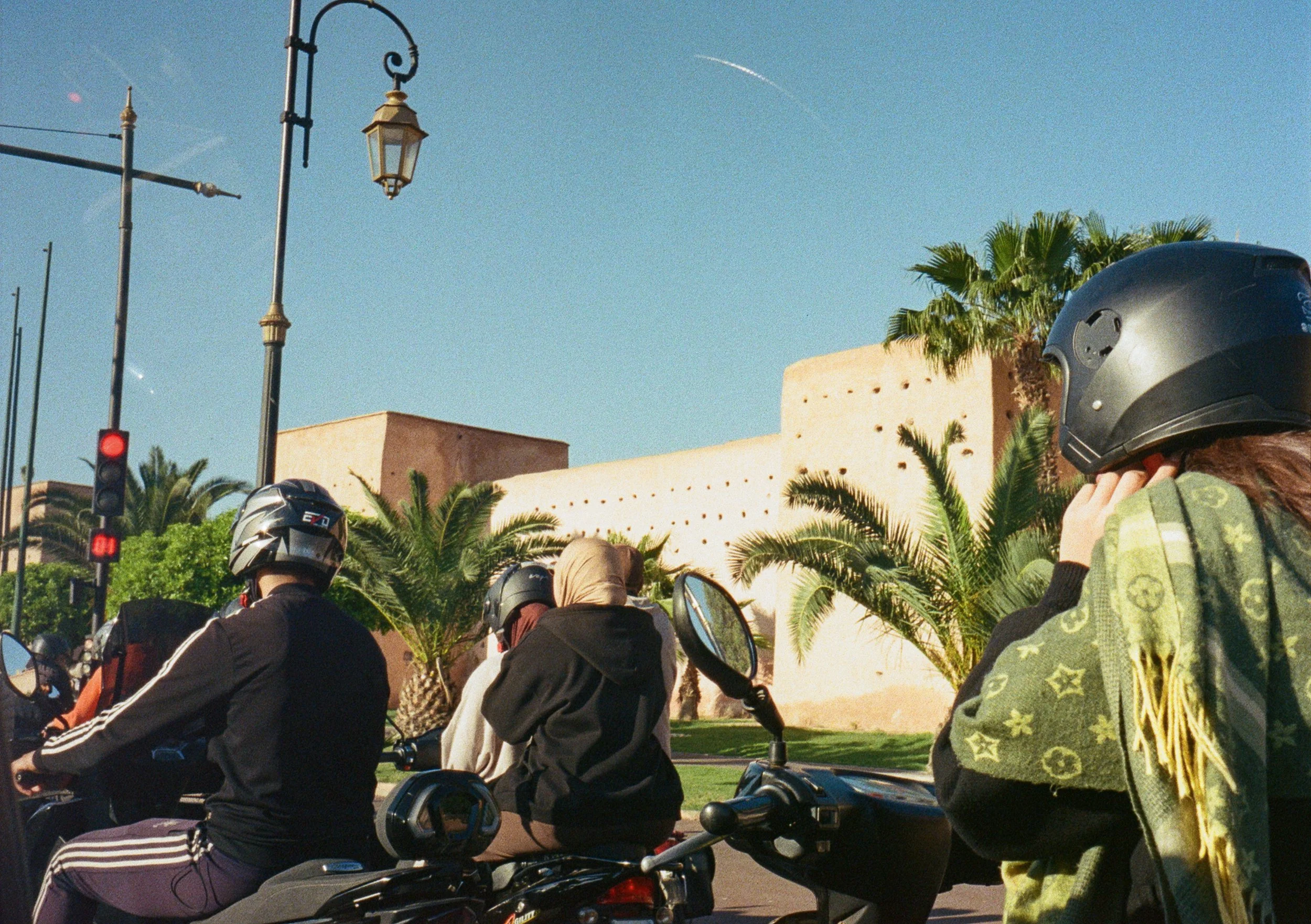 Group of motorcyclists riding on a city street with palm trees, a beige building, street lamps, and a traffic light in the background. One rider is wearing a green patterned hoodie and a black helmet.