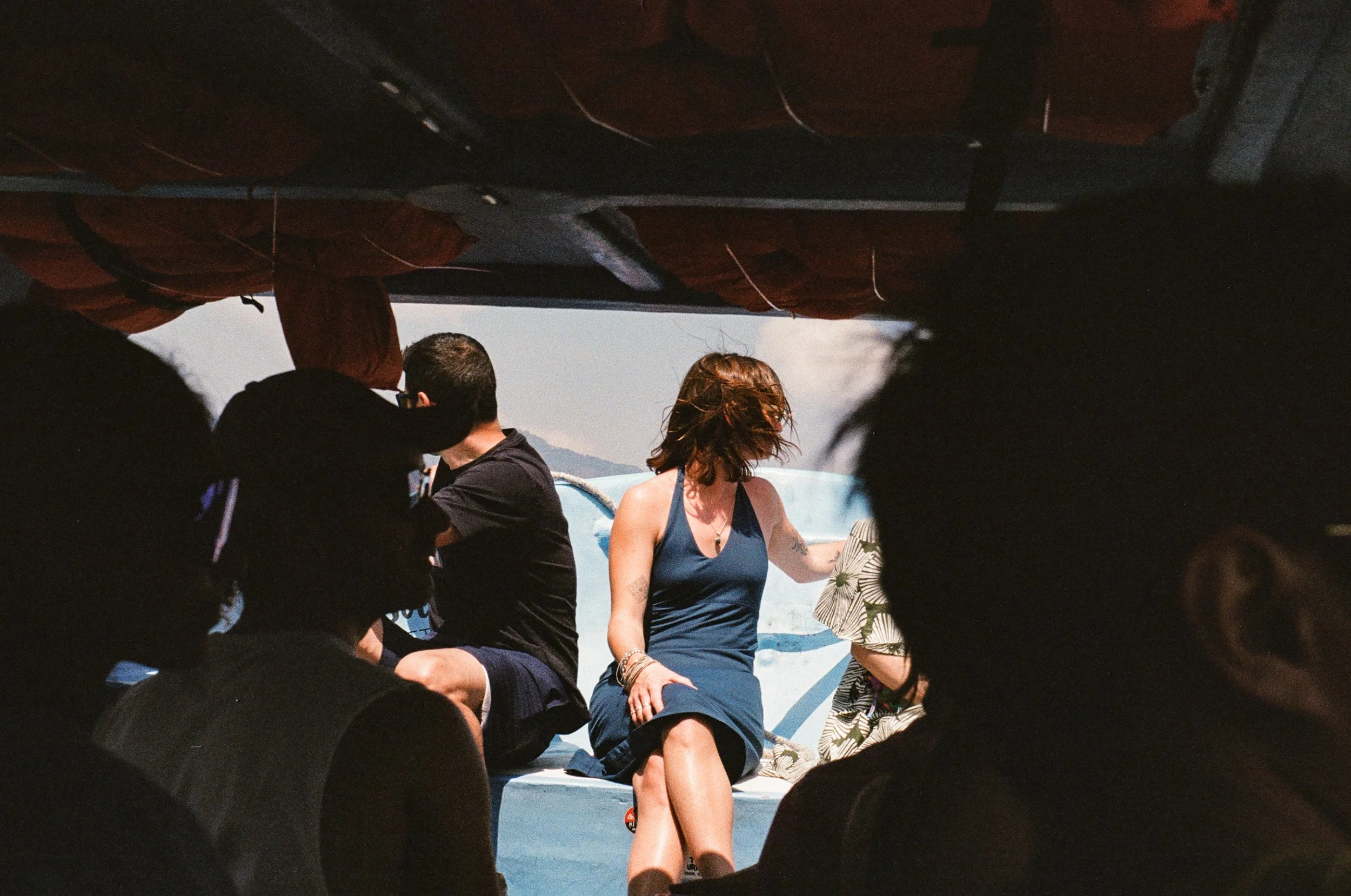 People sitting on a boat, with some standing and some seated, surrounded by water with mountains in the background. The photo is taken from under the boat's canopy.