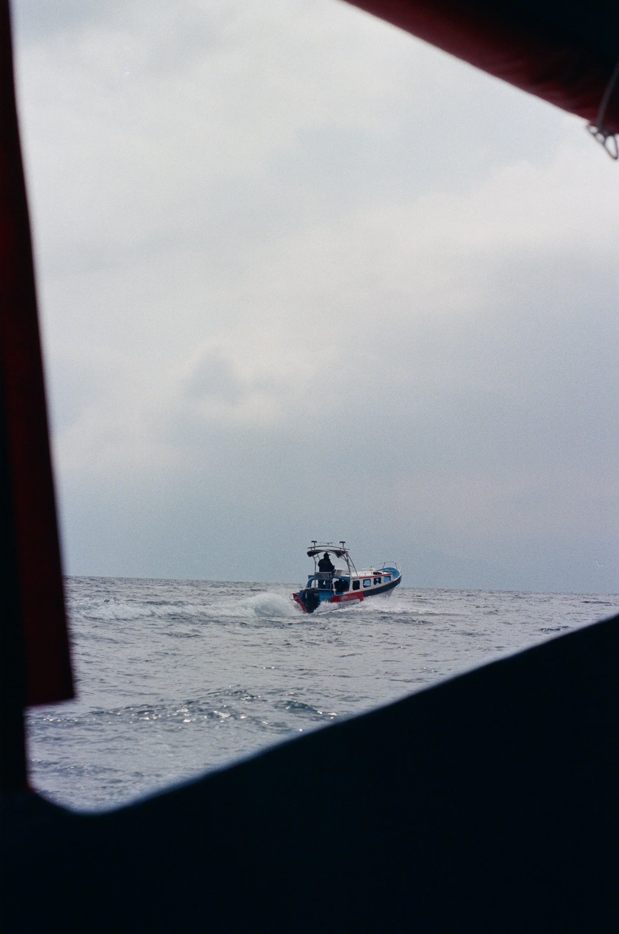 A small boat with a red, white, and blue color scheme is moving through the water, seen through a frame of a window or porthole.