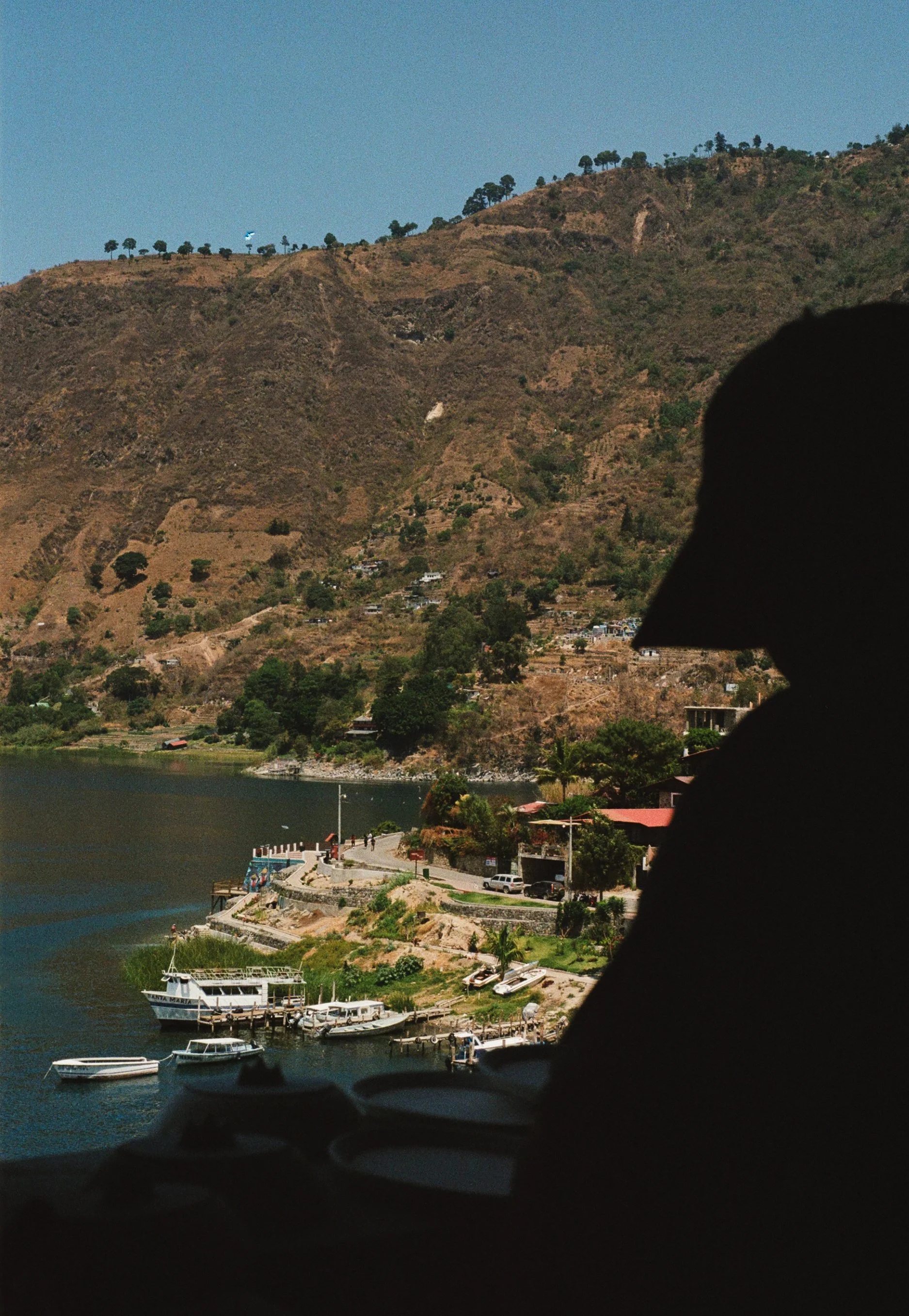 Scenic view of a hillside town near a body of water with boats docked along the shoreline; silhouette of a person's head in the foreground.