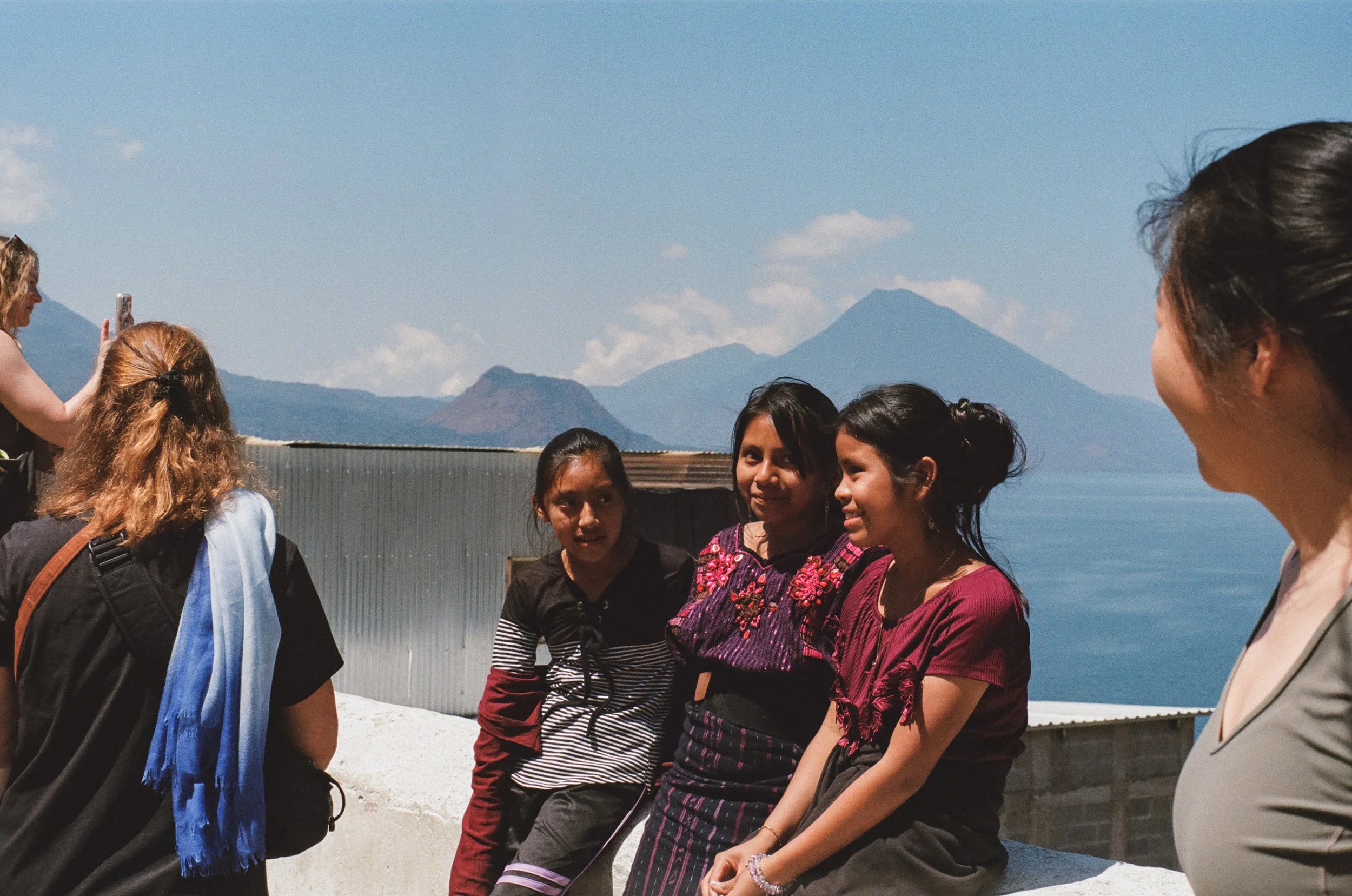 A diverse group of women standing outdoors against a scenic backdrop of mountains, sky, and lake, engaged in conversation and smiling.