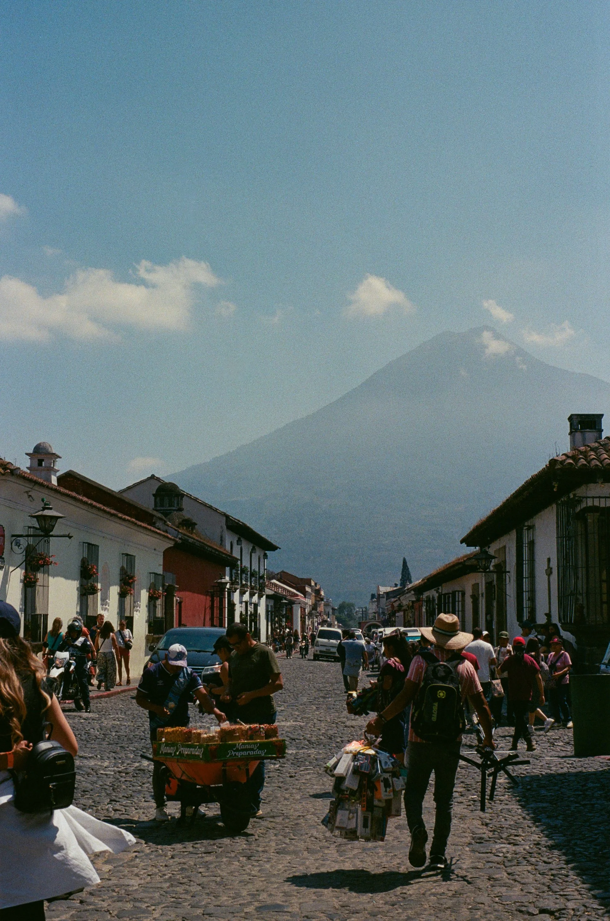 People walking and shopping on a cobblestone street with colorful buildings, mountains in the background, and a clear blue sky.