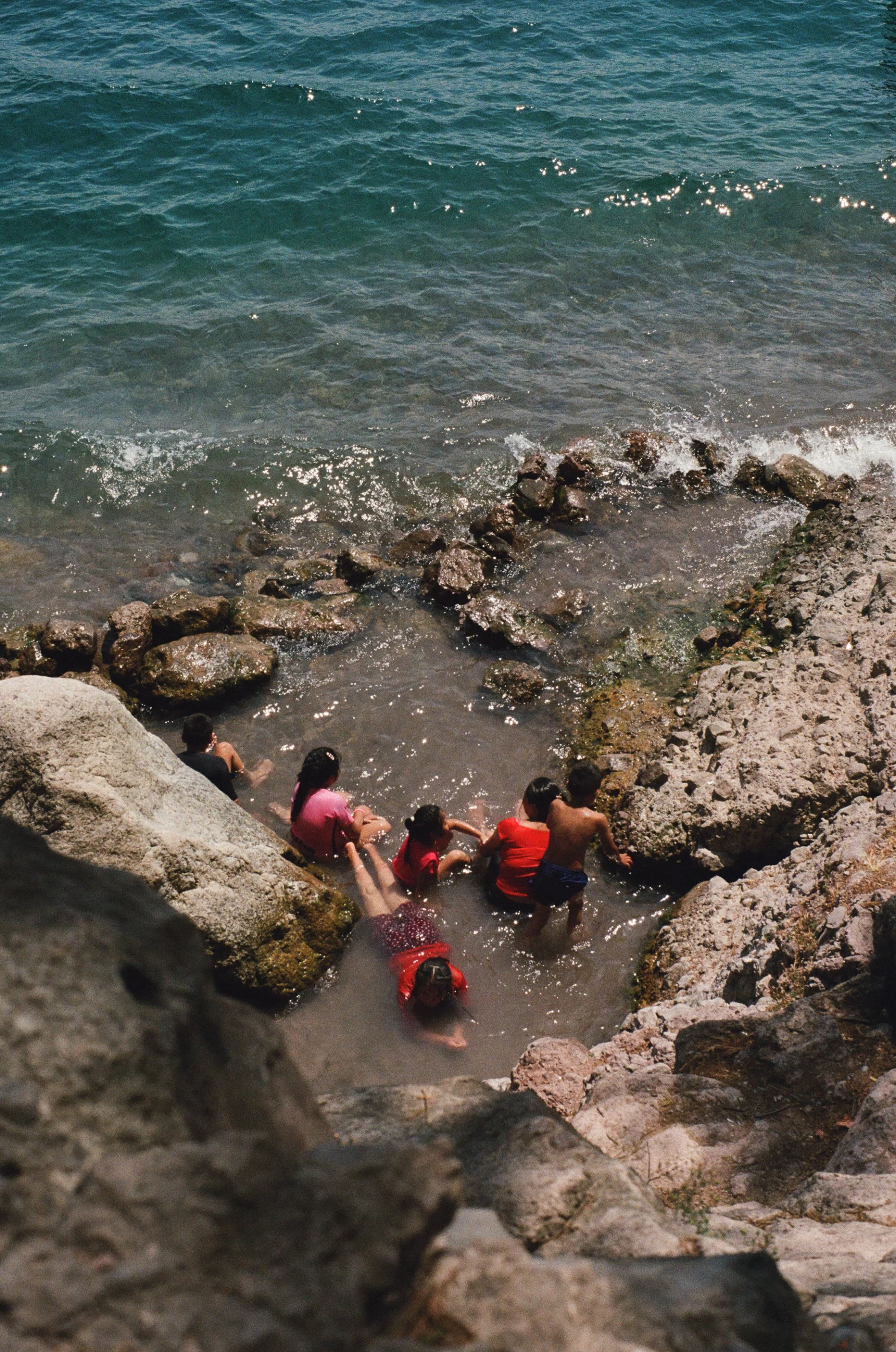People swimming and sitting on rocks at a rocky shoreline by the ocean.