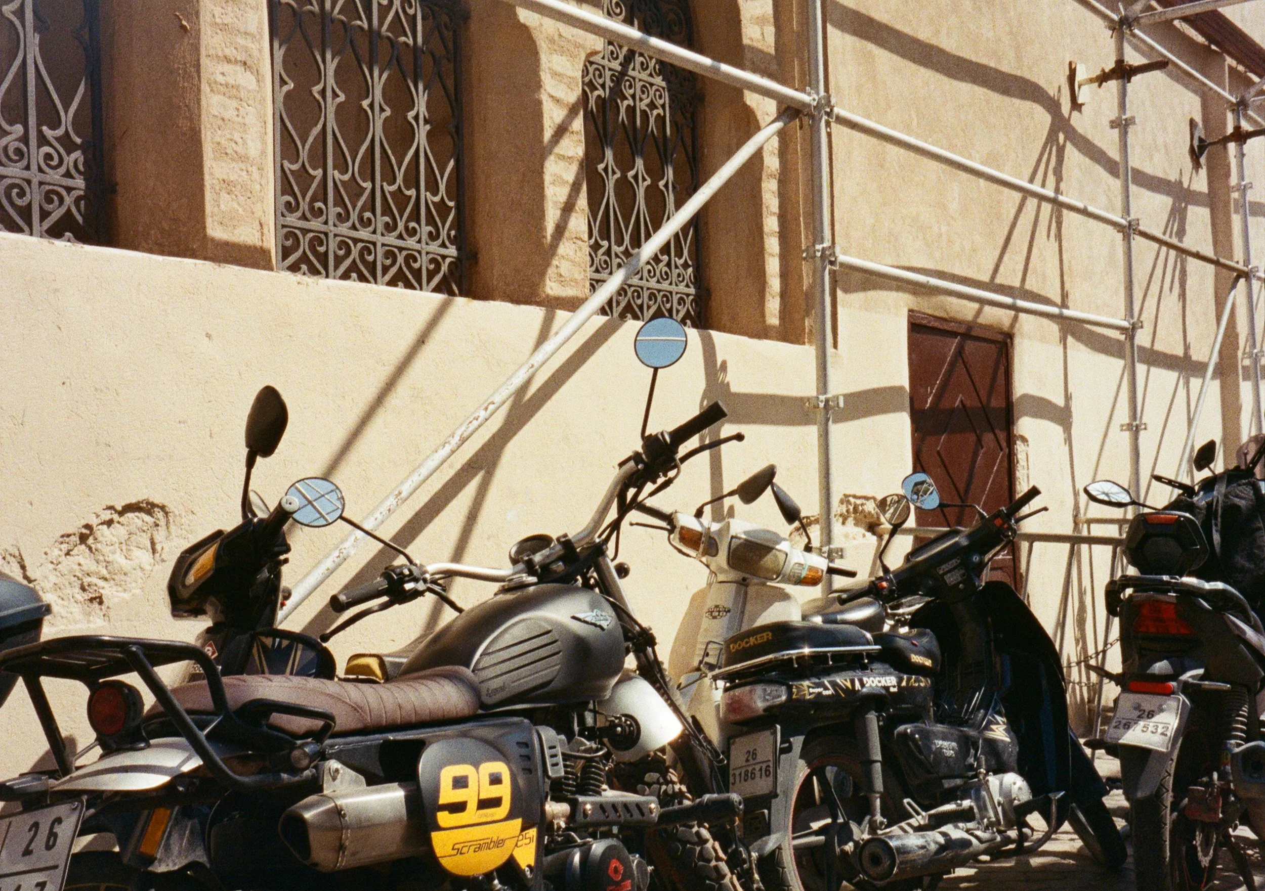 Multiple parked scooters lined up in front of a building under construction, with scaffolding and barred windows.