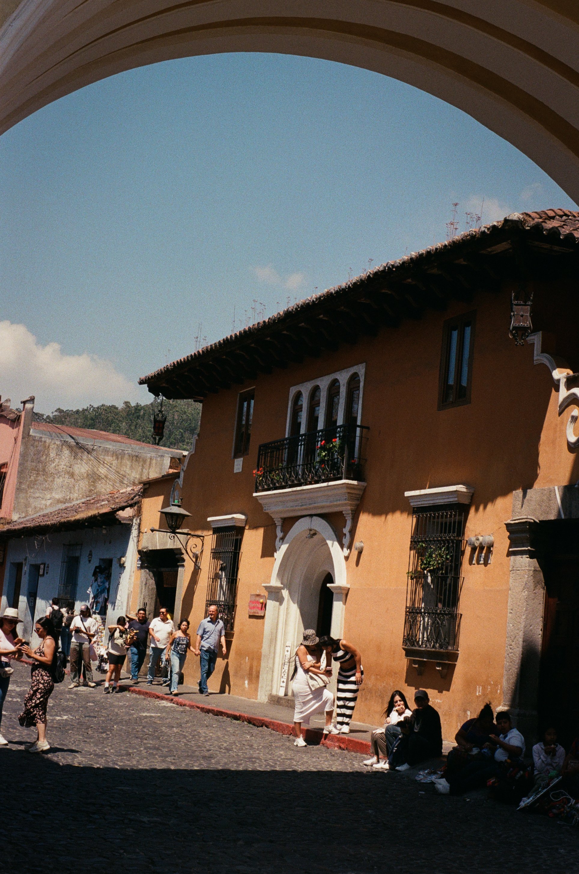 People walking and sitting on a cobblestone street in front of colorful buildings with balconies, under a Spanish-style arch, on a sunny day with a blue sky.
