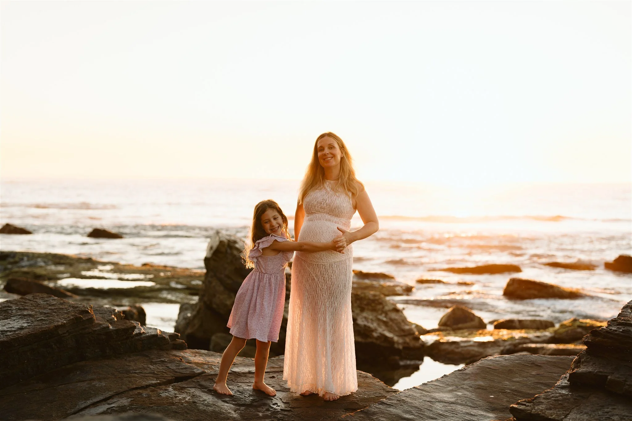 A woman and a young girl standing on rocks by the ocean during sunset, holding hands and smiling.