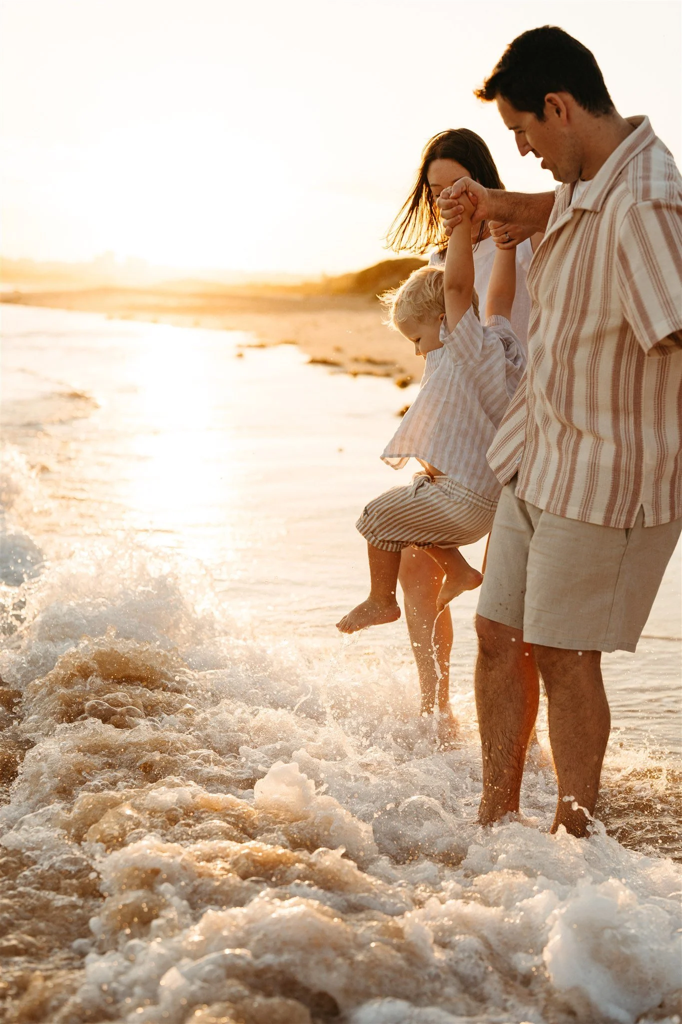 A Mum, Dad and toddler playing on the shoreline in golden light at Long Reef during a golden hour family photography session with Sophie Smith