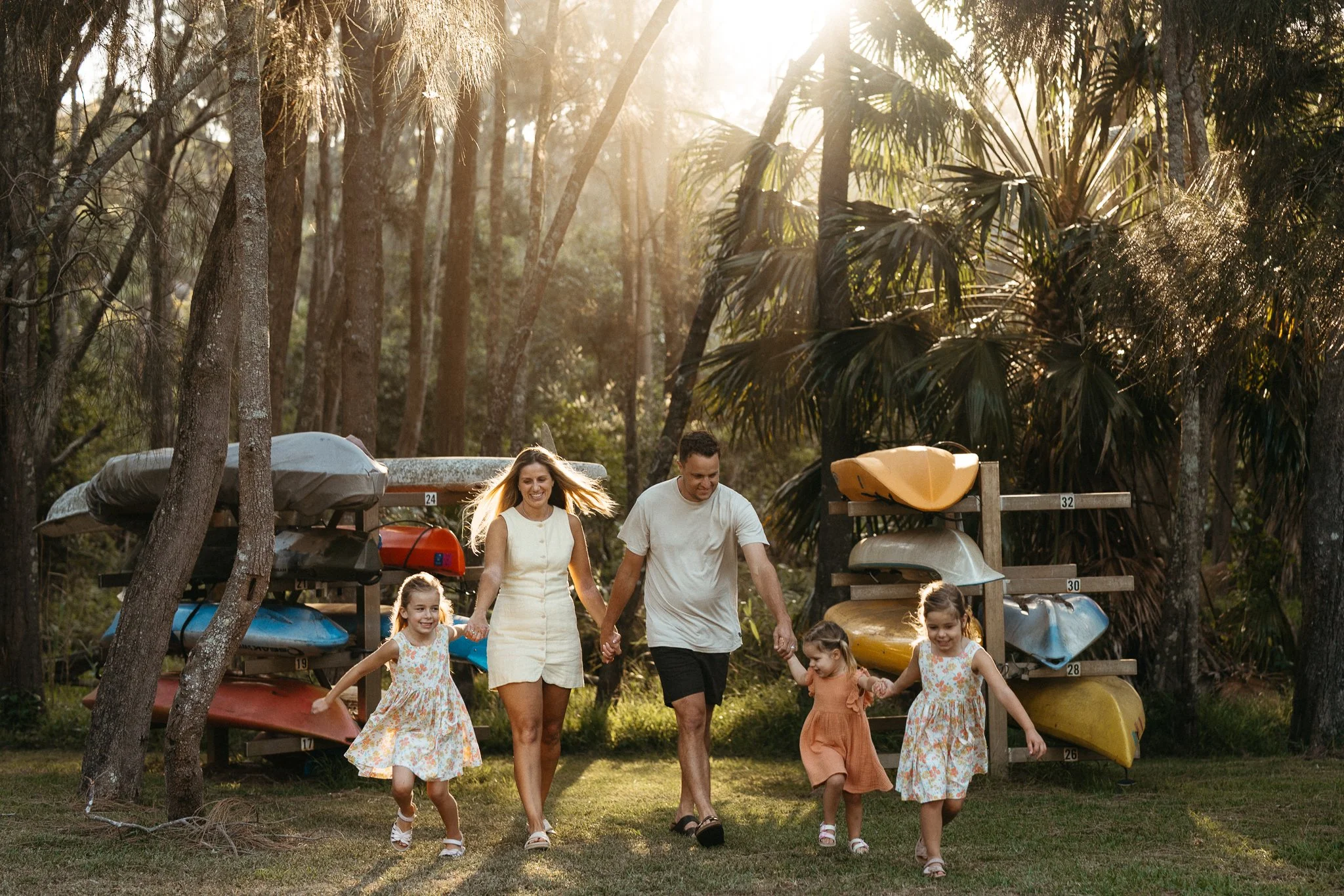 A family with three little girls, running towards the camera with joy on their faces, colourful boats in the background and beautiful golden hour light, near Narrabeen Lake on Sydney's Northern Beaches