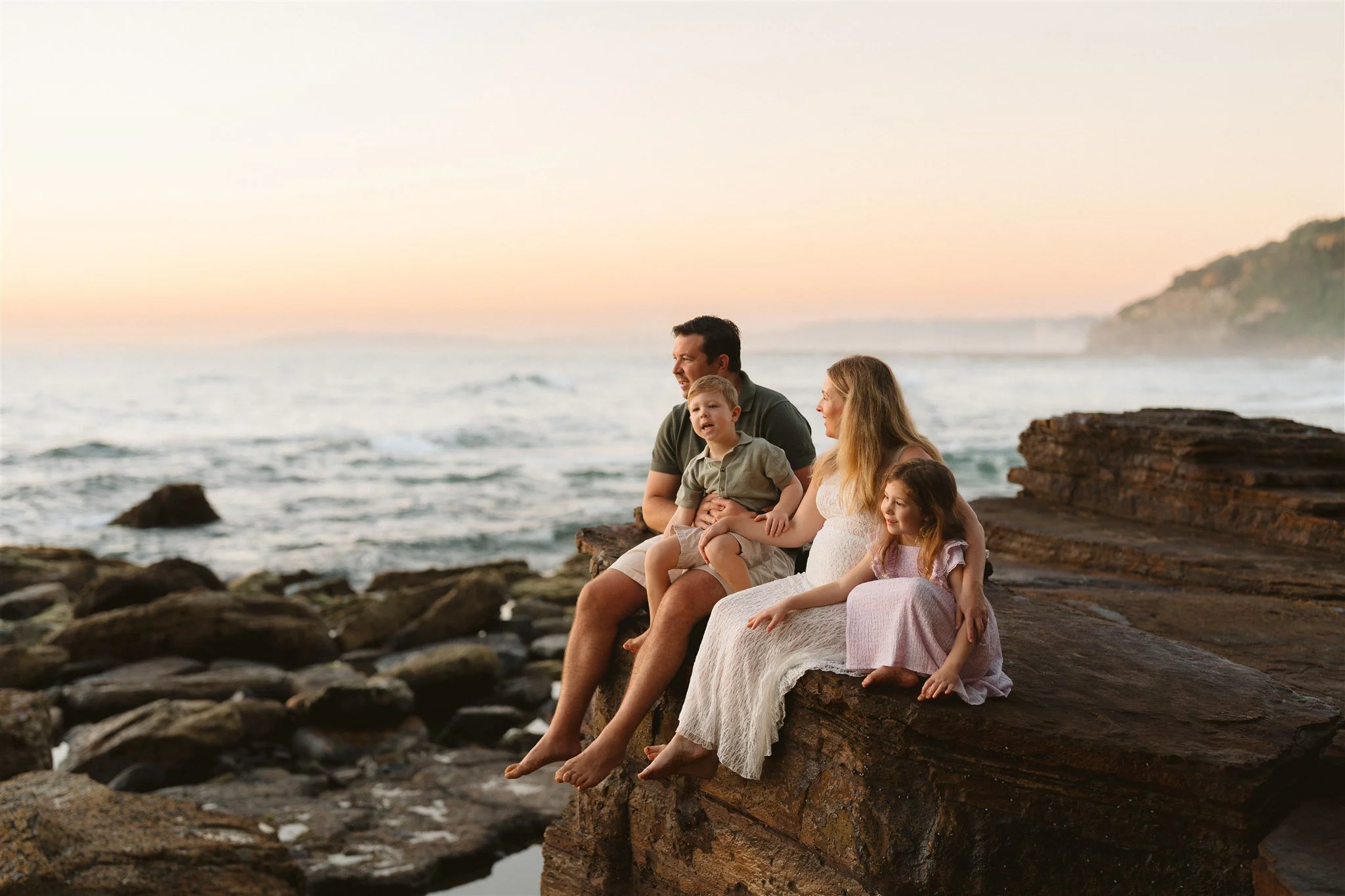 A family of four sitting on a rocky headland at Warriewood Beach looking out towards the ocean during a relaxed family photography session