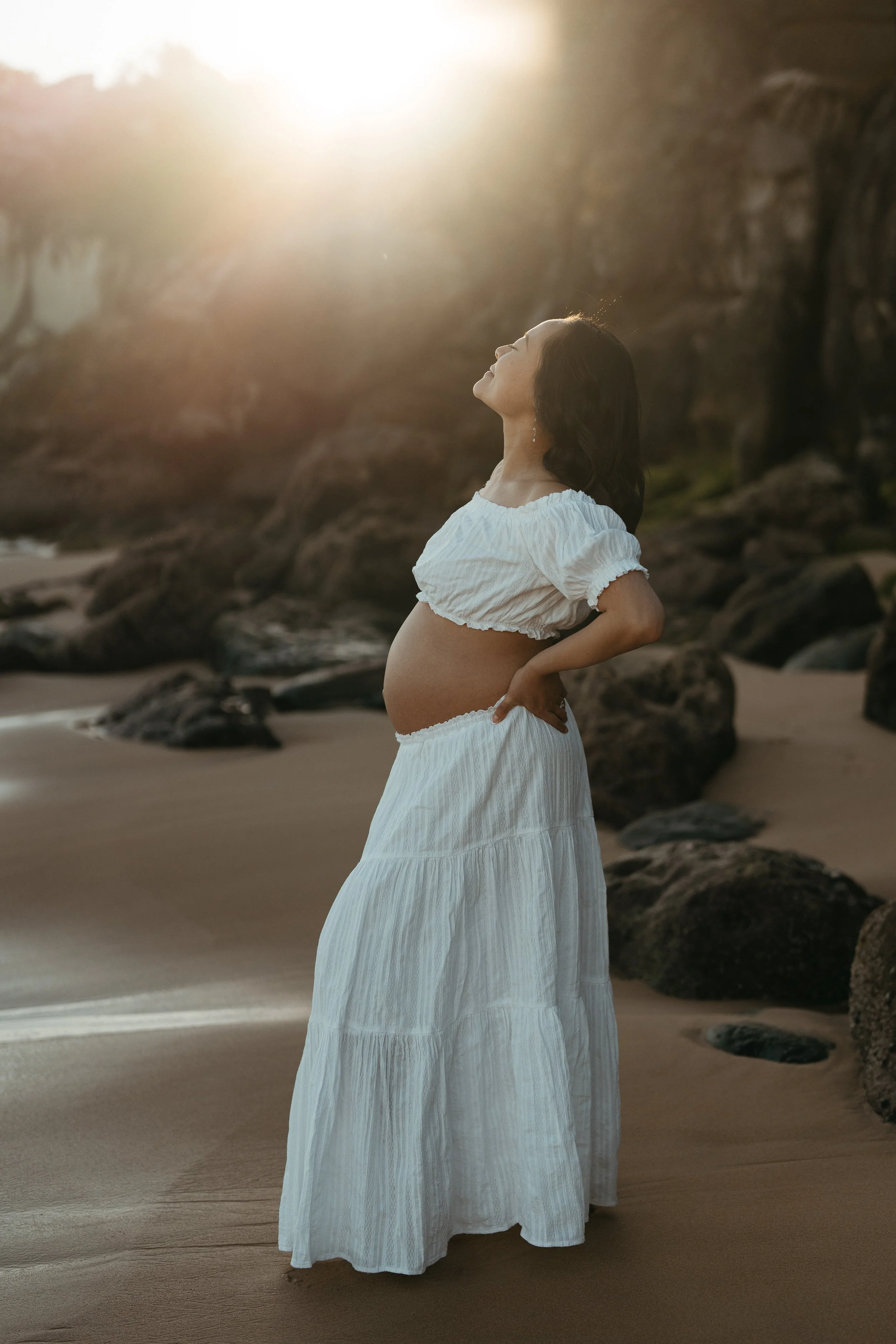 Pregnant woman in white two-piece set, with the sun setting behind her, during a maternity photography session at Curl Curl beach in Sydney
